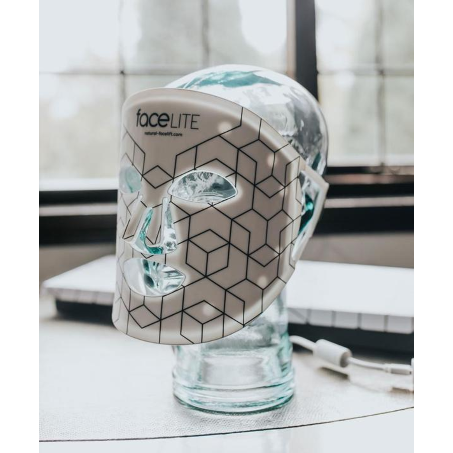 A FaceLite mask on a glass head. The mask is white with black geometric patterns. The background is blurred.