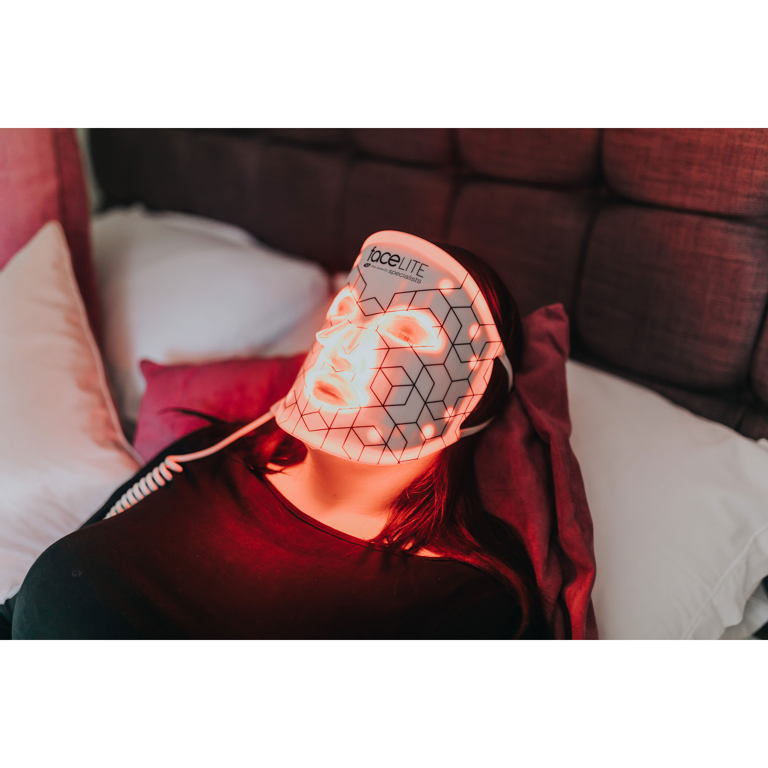 A woman wearing a face mask with red light therapy, lying on a bed with pillows.