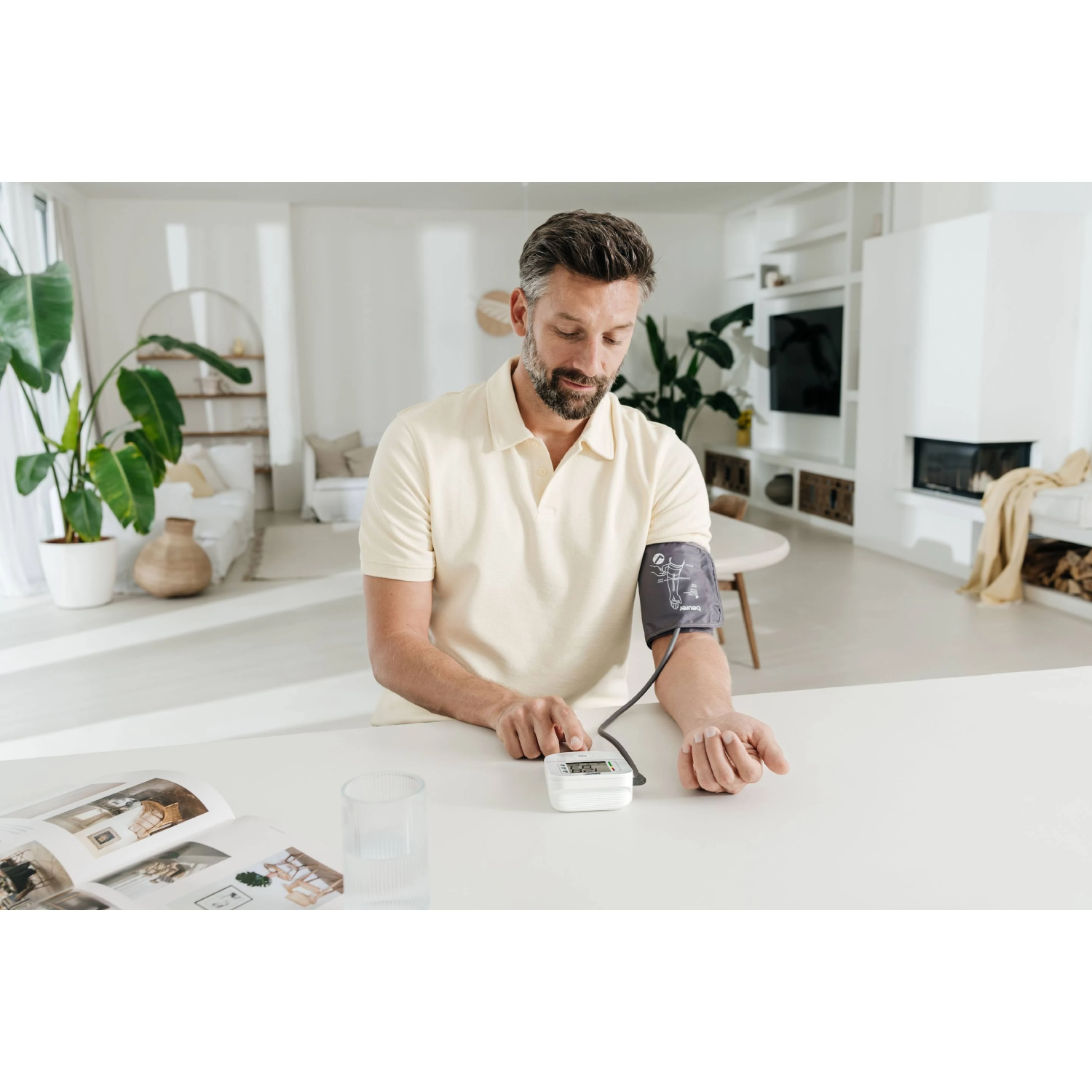 Un hombre se toma la tensión con un monitor digital. Está en un salón moderno y luminoso. En la mesa hay una revista y un vaso.