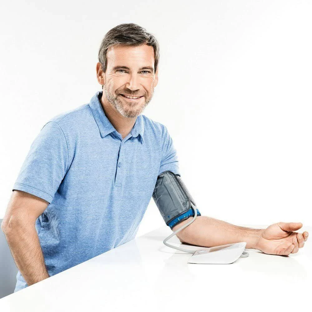 Un hombre de pelo gris sonríe. Tiene un monitor de presión arterial en el brazo. Está sentado en una mesa blanca.