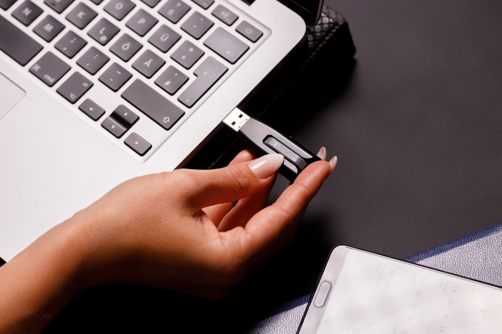 Bir el, bir USB sürücüsünü bir dizüstü bilgisayara takıyor. Arka planda bir akıllı telefon ve not defteri siyah bir yüzeyde duruyor.
