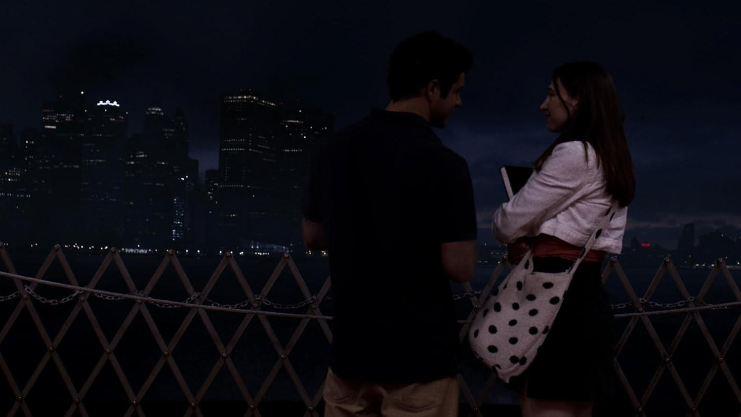 Zwei Personen auf einer Brücke bei Nacht, Skyline im Hintergrund. Eine Person hält eine Polka-Dot-Tasche.
