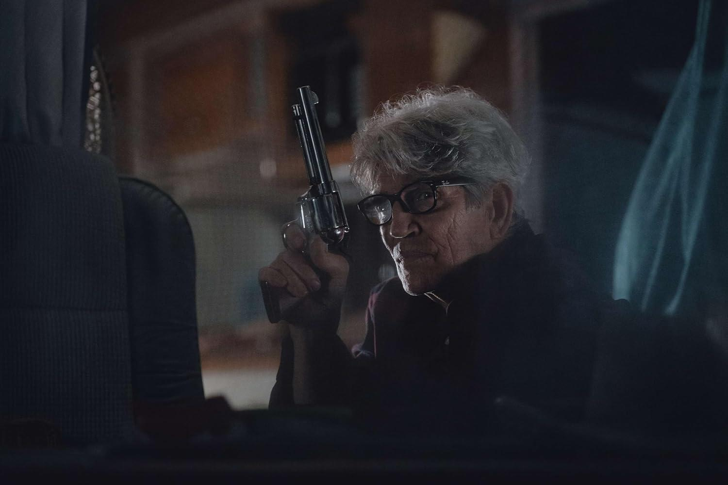Ein älterer Mann mit Brille hält eine Waffe. Er ist in einem Auto. Der Hintergrund ist dunkel.