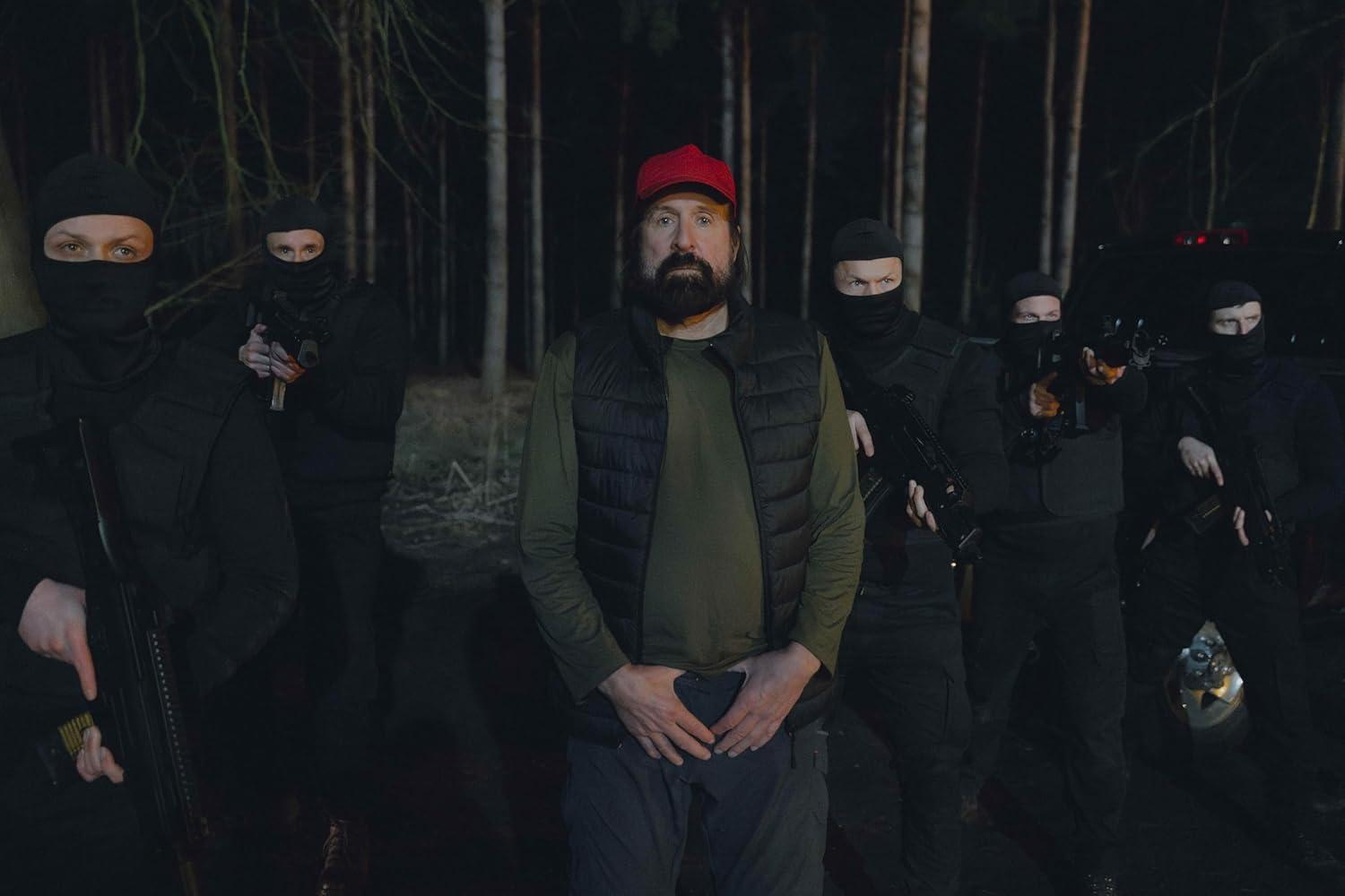 Ein Mann mit roter Mütze und Bart steht mit maskierten Gestalten, die Waffen halten, in einem Wald bei Nacht.