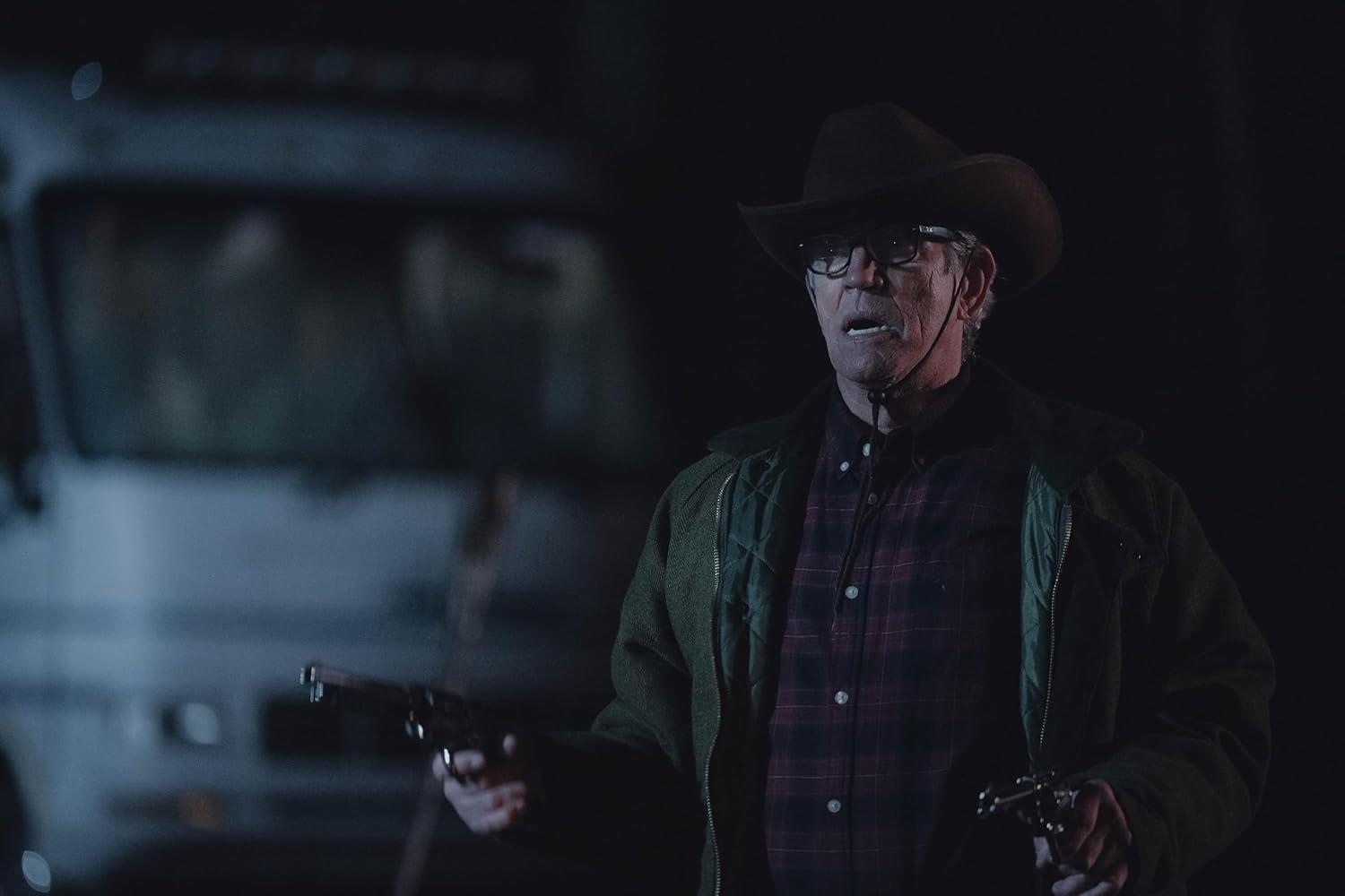 Mann mit Cowboyhut hält zwei Pistolen. Er trägt eine Brille, ein kariertes Hemd und eine grüne Jacke. Ein Van ist unscharf im Hintergrund.