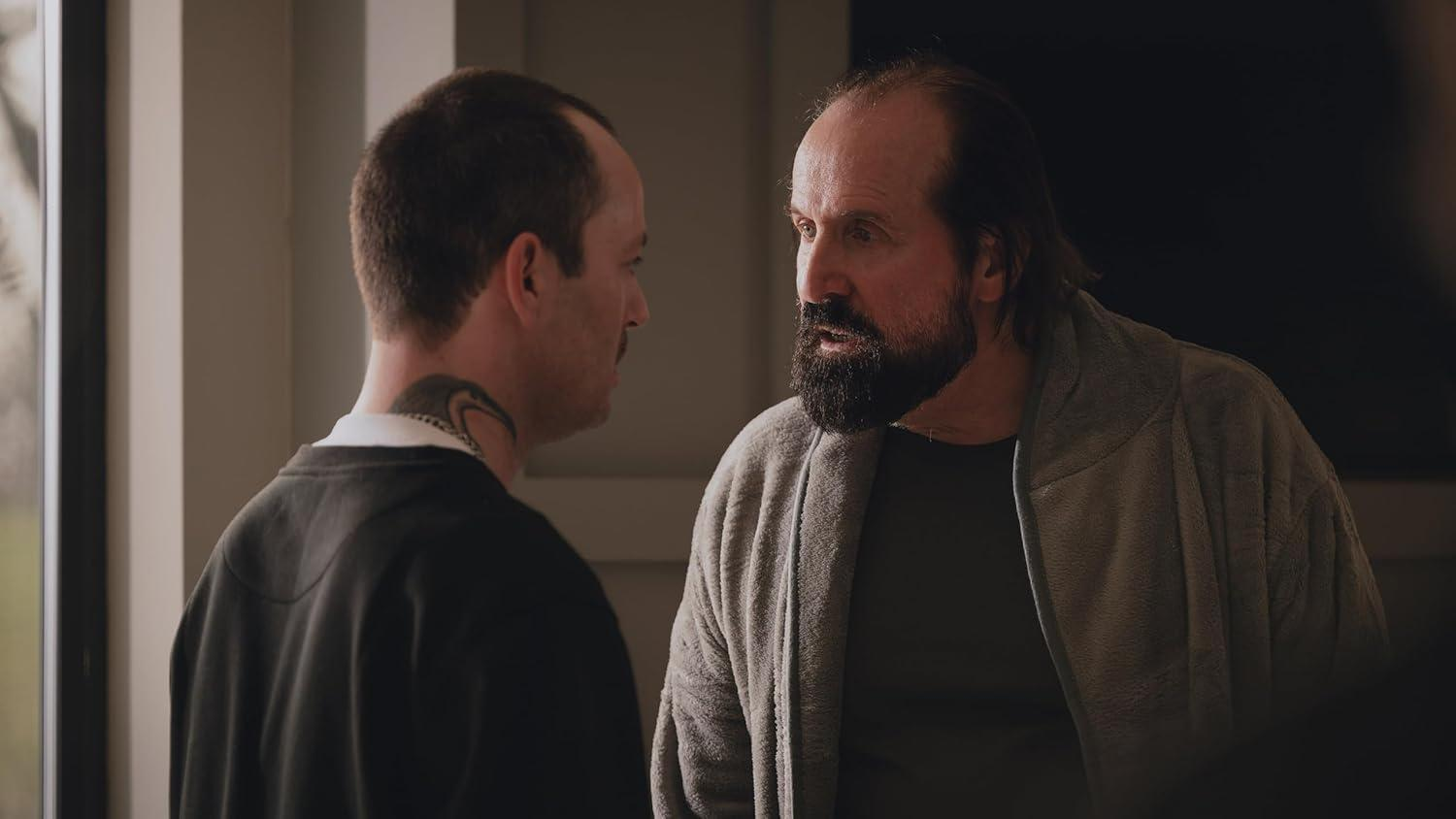 Zwei Männer in einem Raum. Einer mit Bart und Bademantel, der andere mit einem Hals-Tattoo. Sie schauen sich an und reden.