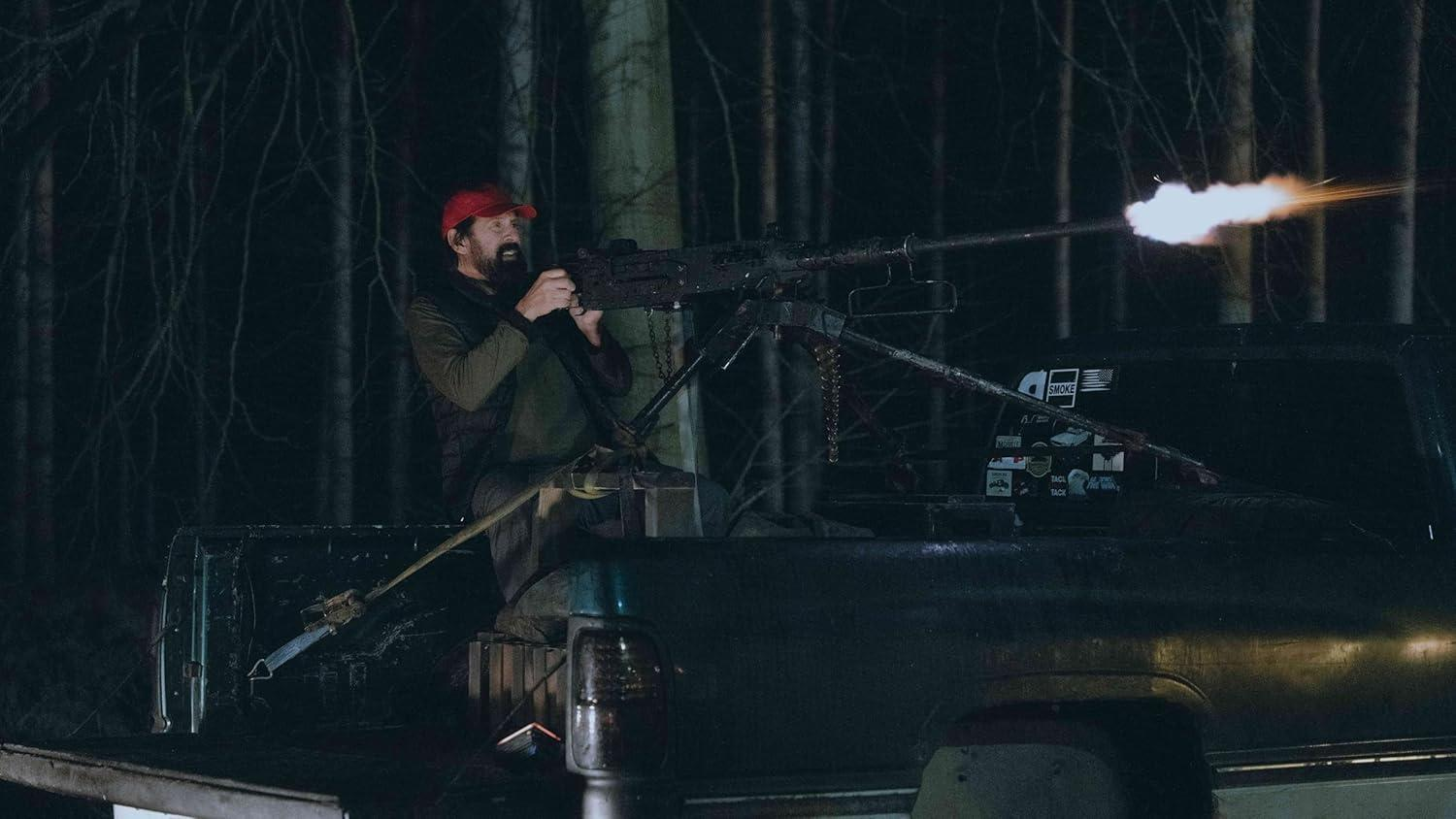 Mann mit roter Kappe feuert aus einer Ladefläche heraus mit einem Maschinengewehr in die Nacht. Heller Mündungsblitz erhellt die Szene. Wald im Hintergrund.