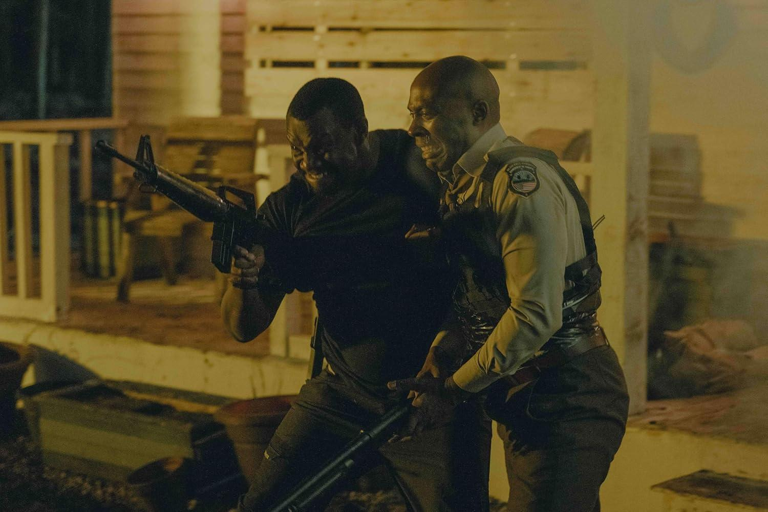 Zwei Männer, einer mit einem Gewehr, scheinen sich in einer angespannten Situation zu befinden. Im Hintergrund ist ein Gebäude bei Nacht zu sehen.