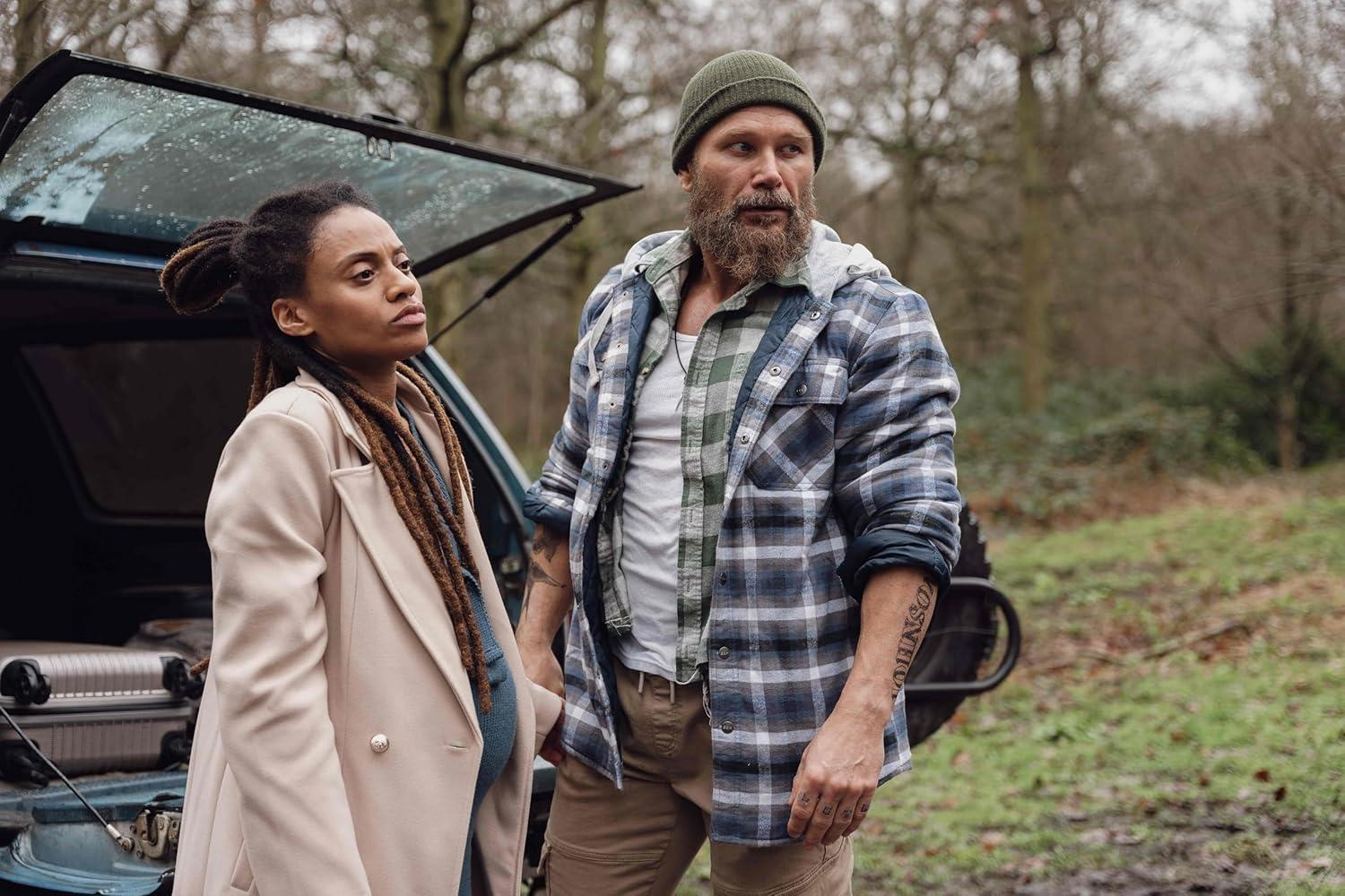 Eine Frau mit Dreadlocks und ein Mann mit Bart stehen in einem Wald an einem Auto. Der Mann trägt ein kariertes Hemd und eine Mütze.