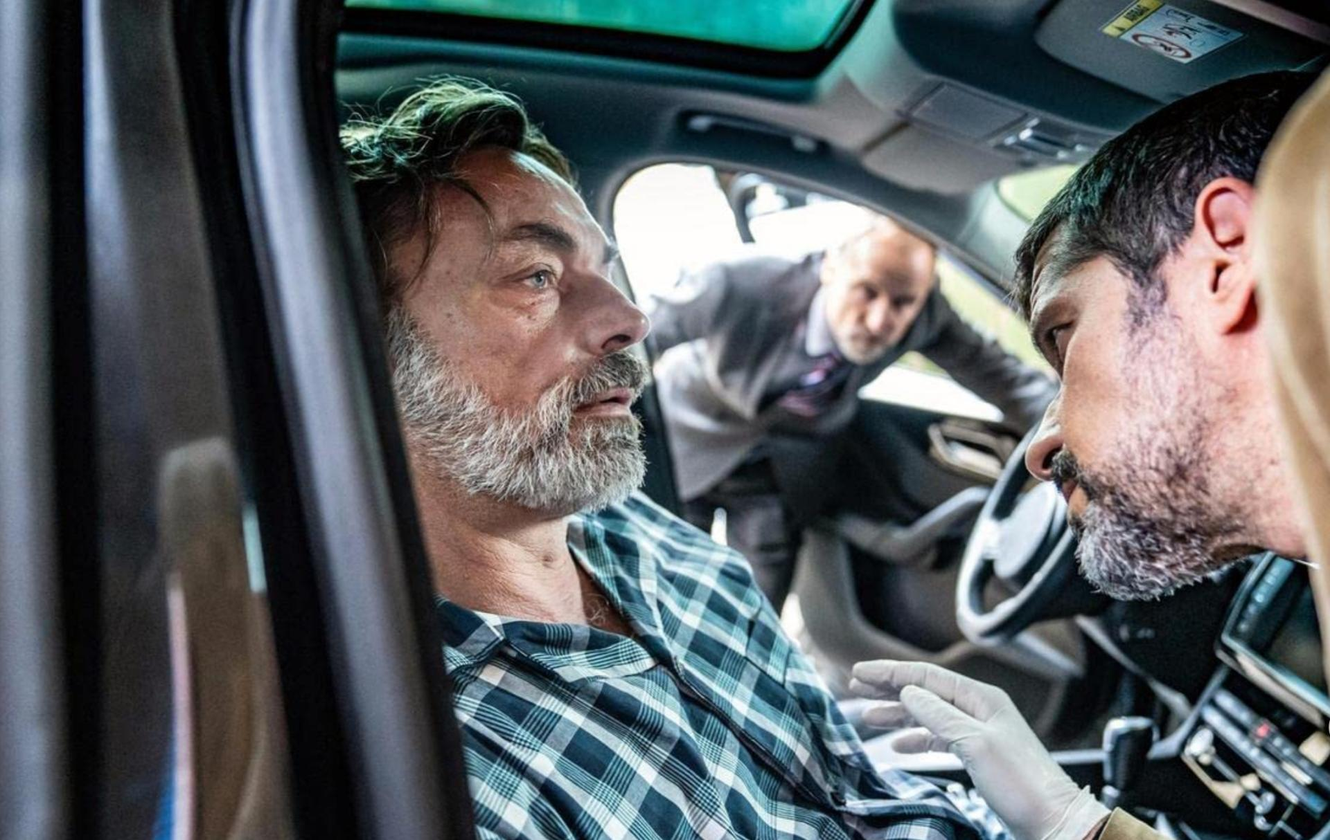 Ein Mann im Auto blickt besorgt, als sich ein anderer Mann im Anzug nähert. Ein dritter Mann in Handschuhen lehnt sich vor.