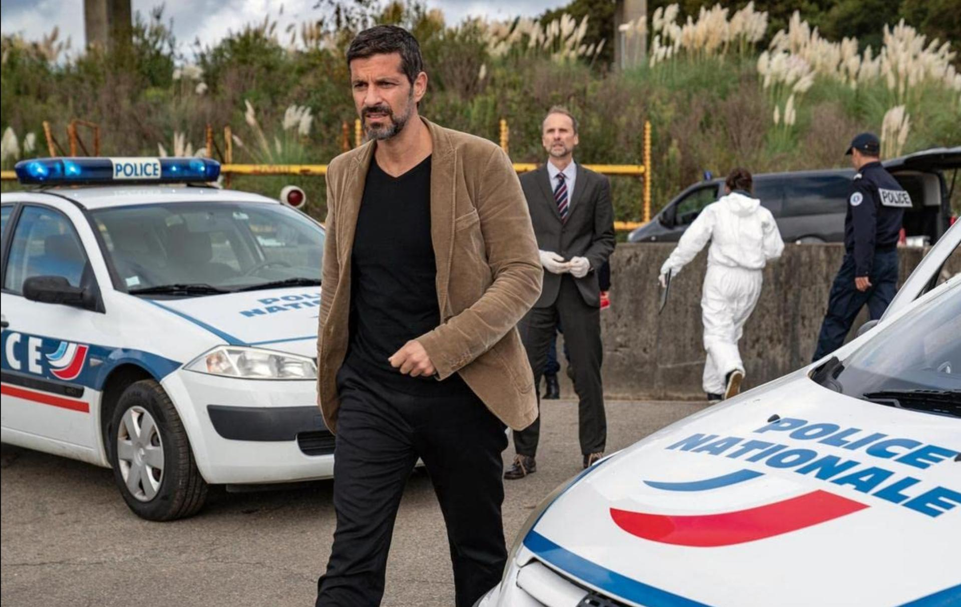 Ein Mann in brauner Jacke geht an Polizeiautos an einem Tatort vorbei. Andere Leute in Anzügen und weißem Anzug sind da.