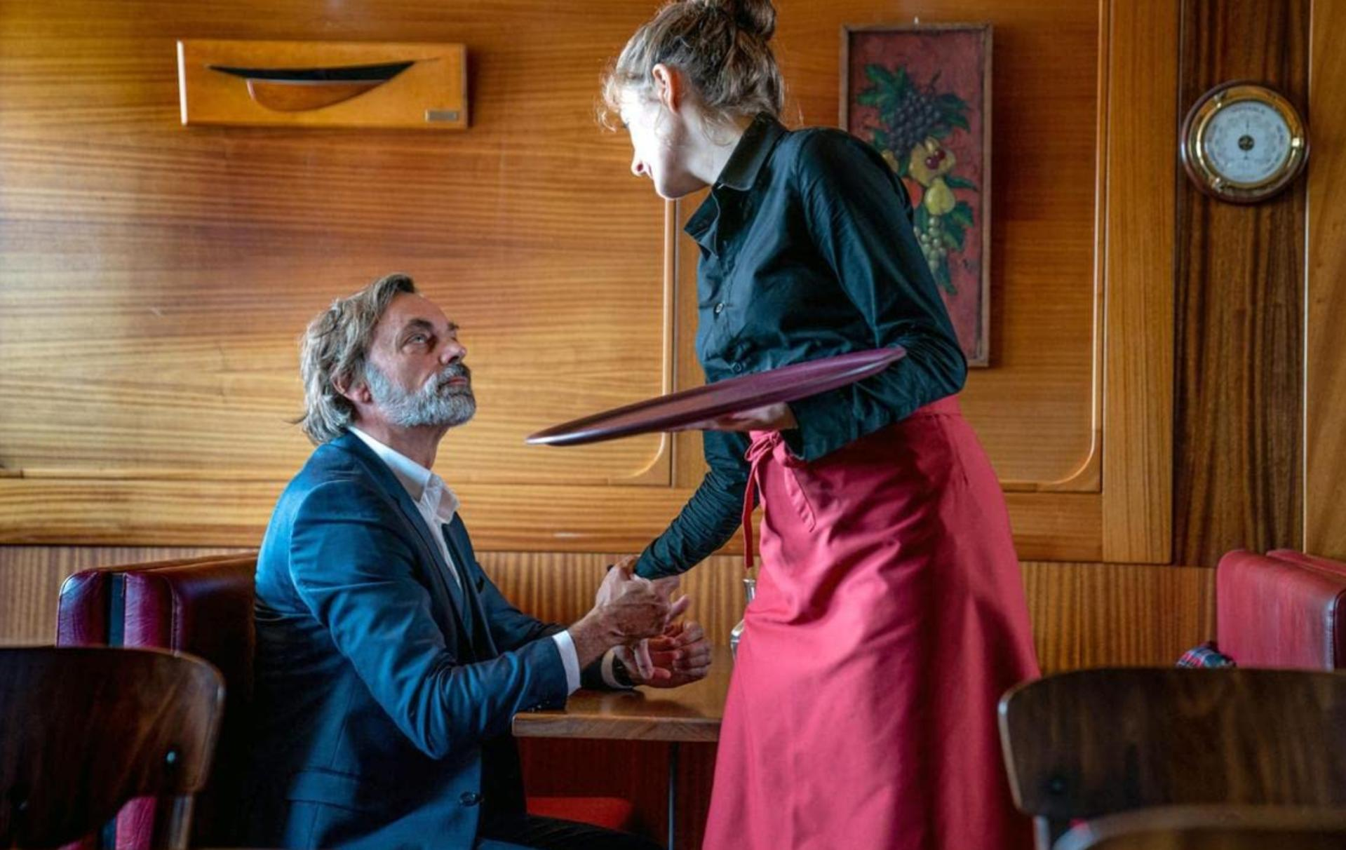 Eine Kellnerin bietet einem Mann im blauen Anzug ein Tablett an. Der Mann sitzt in einem Restaurant an einem Tisch.
