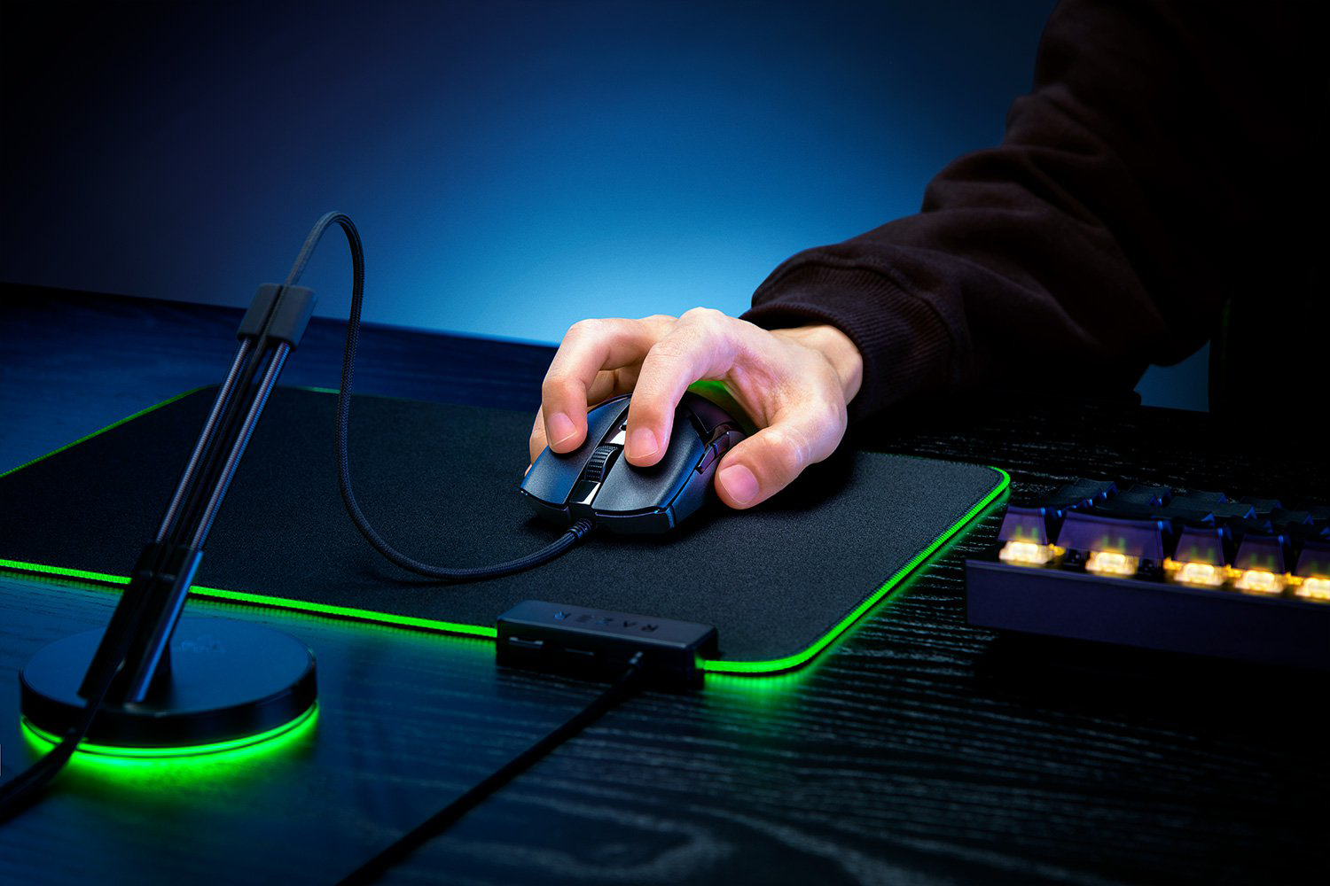 Egy kéz egy fekete gamer egeret használ egy fekete egérpadon, zöld szegélyvilágítással. Egy billentyűzet is látható.