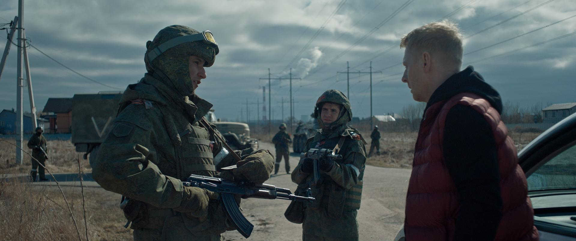 Soldaten in Tarnuniformen mit Gewehren unterhalten sich mit einem Mann in roter Weste. Ein Militärfahrzeug ist im Hintergrund.