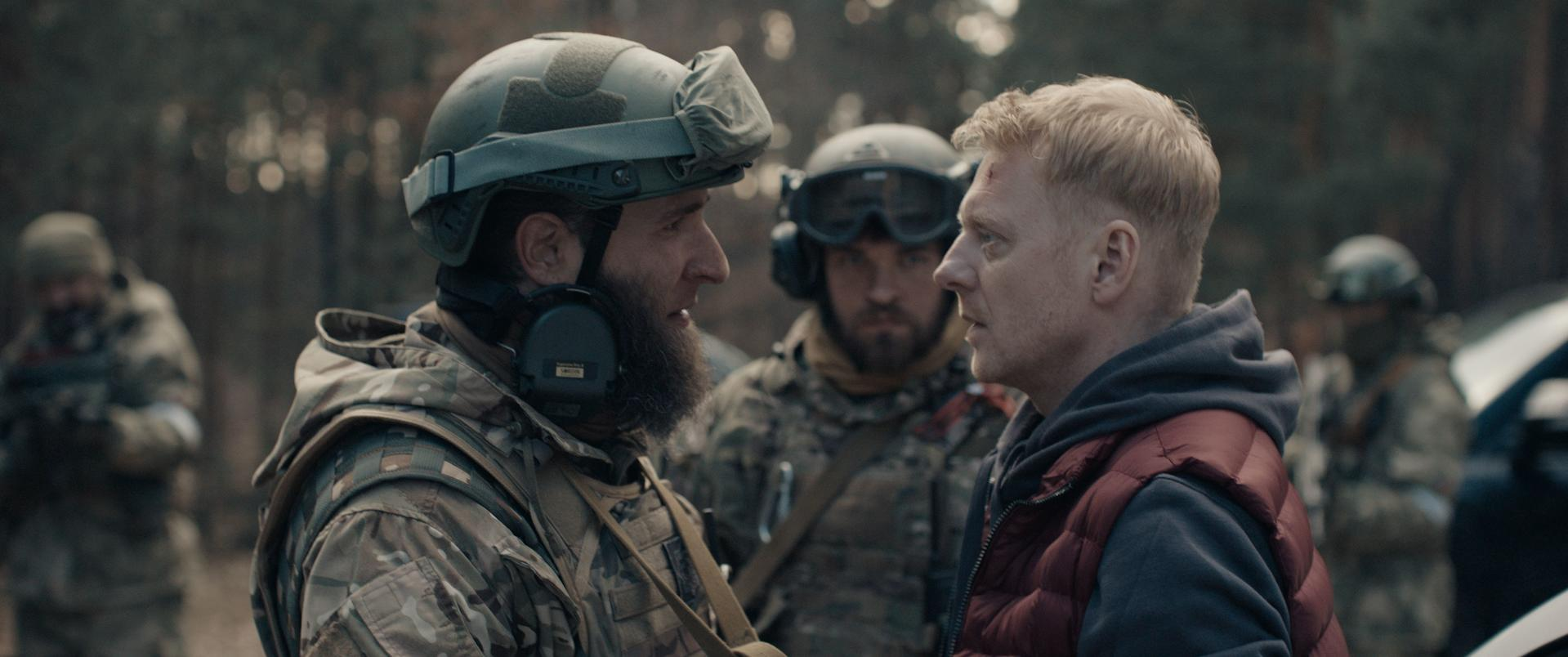 Soldat in Tarnung, Helm und Bart, blickt einen Mann in einer roten Weste an. Andere Soldaten und ein Wald sind im Hintergrund.