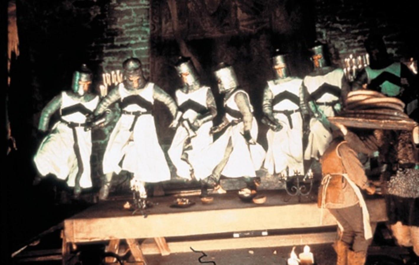 Sechs Ritter in weißen Roben mit schwarzen Kreuzen stehen auf einem Tisch. Ein Mann in brauner Kleidung steht daneben, mit Kerzen und Geschirr.