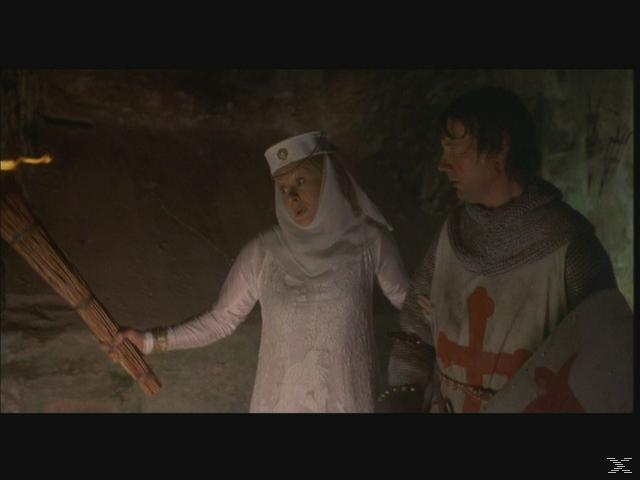 Eine Frau in Weiß hält eine Fackel, neben einem Ritter in Rüstung. Sie sind in einer schwach beleuchteten Höhle. Ein rotes Kreuz ist auf dem Schild des Ritters.