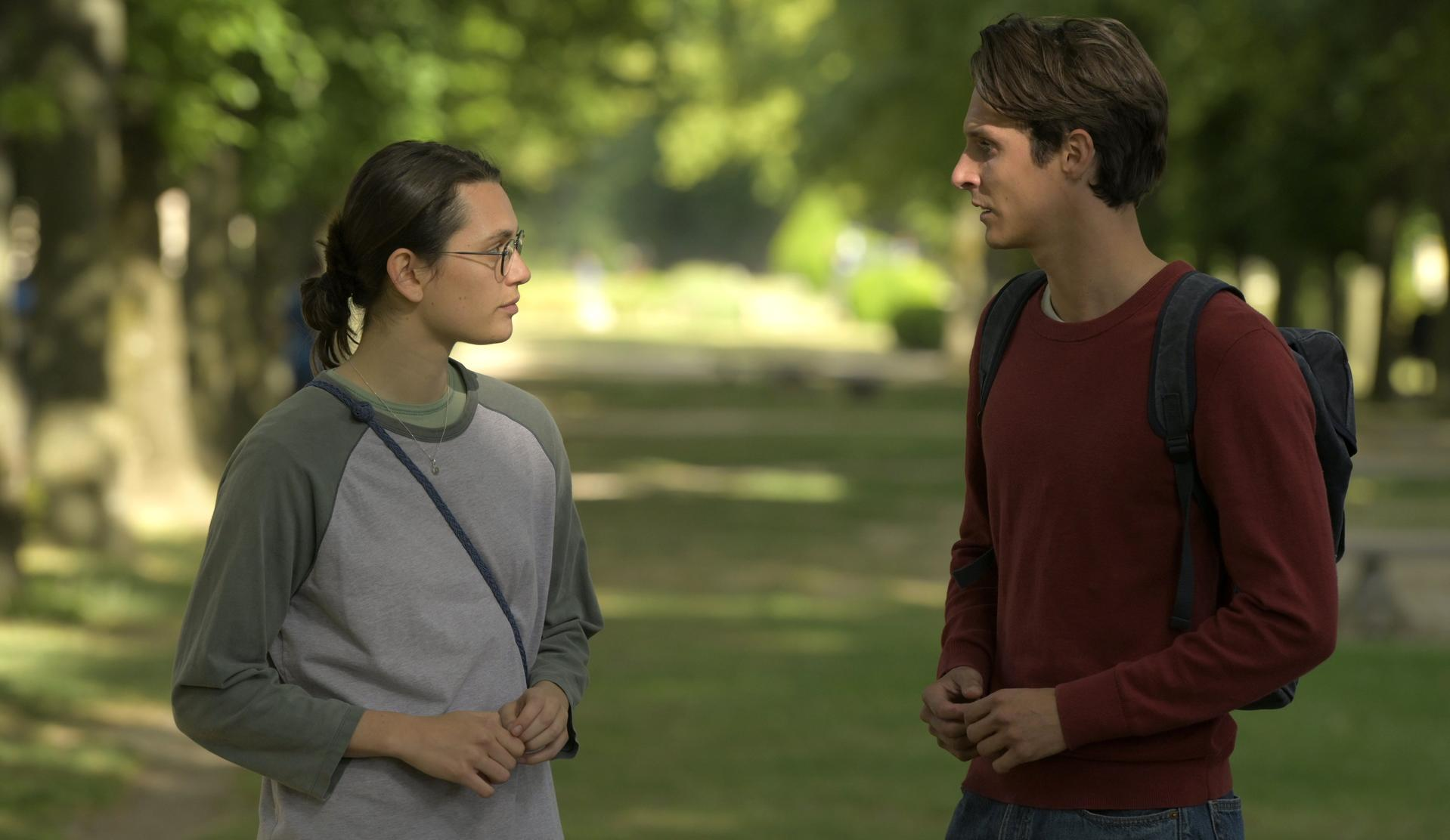 Zwei Personen stehen sich in einem Park gegenüber. Einer trägt eine Brille und ein graues Hemd, der andere einen roten Pullover mit Rucksack.