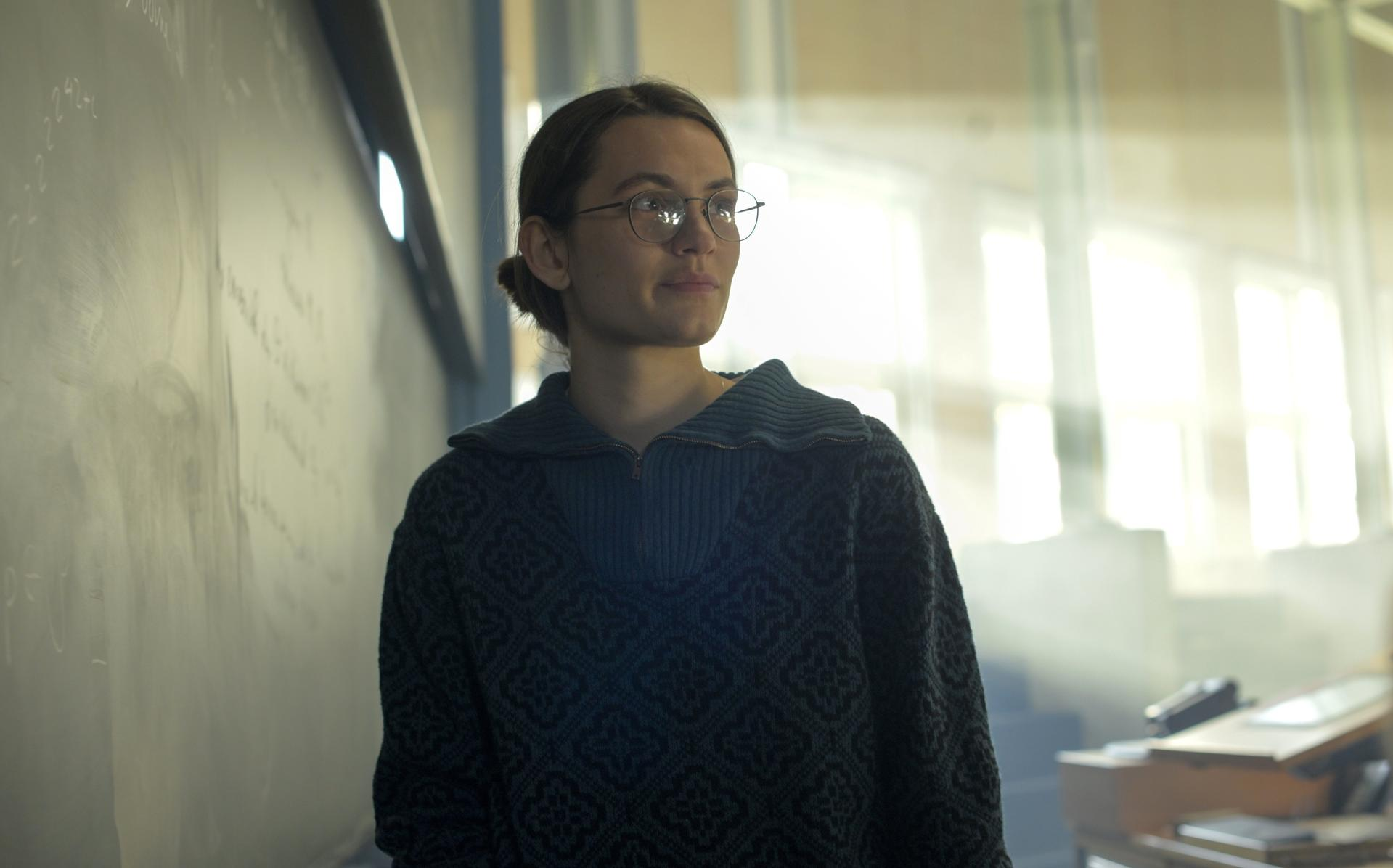 Eine junge Frau mit Brille in einem Klassenzimmer. Sie blickt in Richtung Licht, eine Tafel hinter ihr. Sie trägt einen dunklen Pullover.