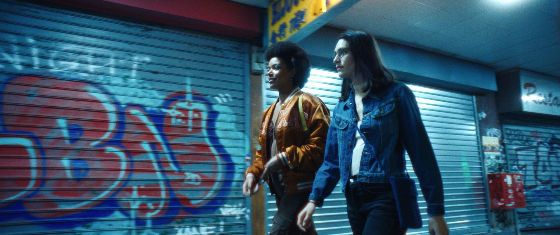 Zwei Frauen gehen an mit Graffiti bedeckten Schaufenstern vorbei. Eine trägt eine Jacke, die andere eine Jeansjacke. Die Szene hat einen blauen Farbton.