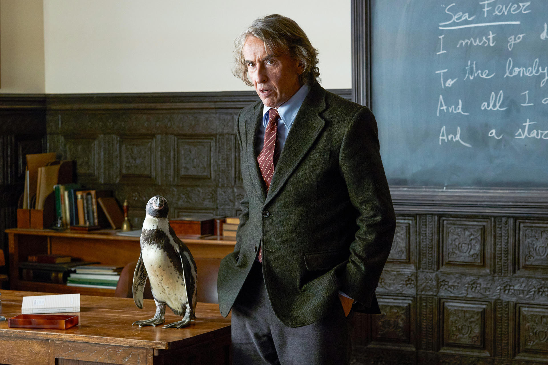 Ein Mann im Anzug steht neben einem Pinguin auf einem Schreibtisch. Im Hintergrund befindet sich eine Tafel mit Text.