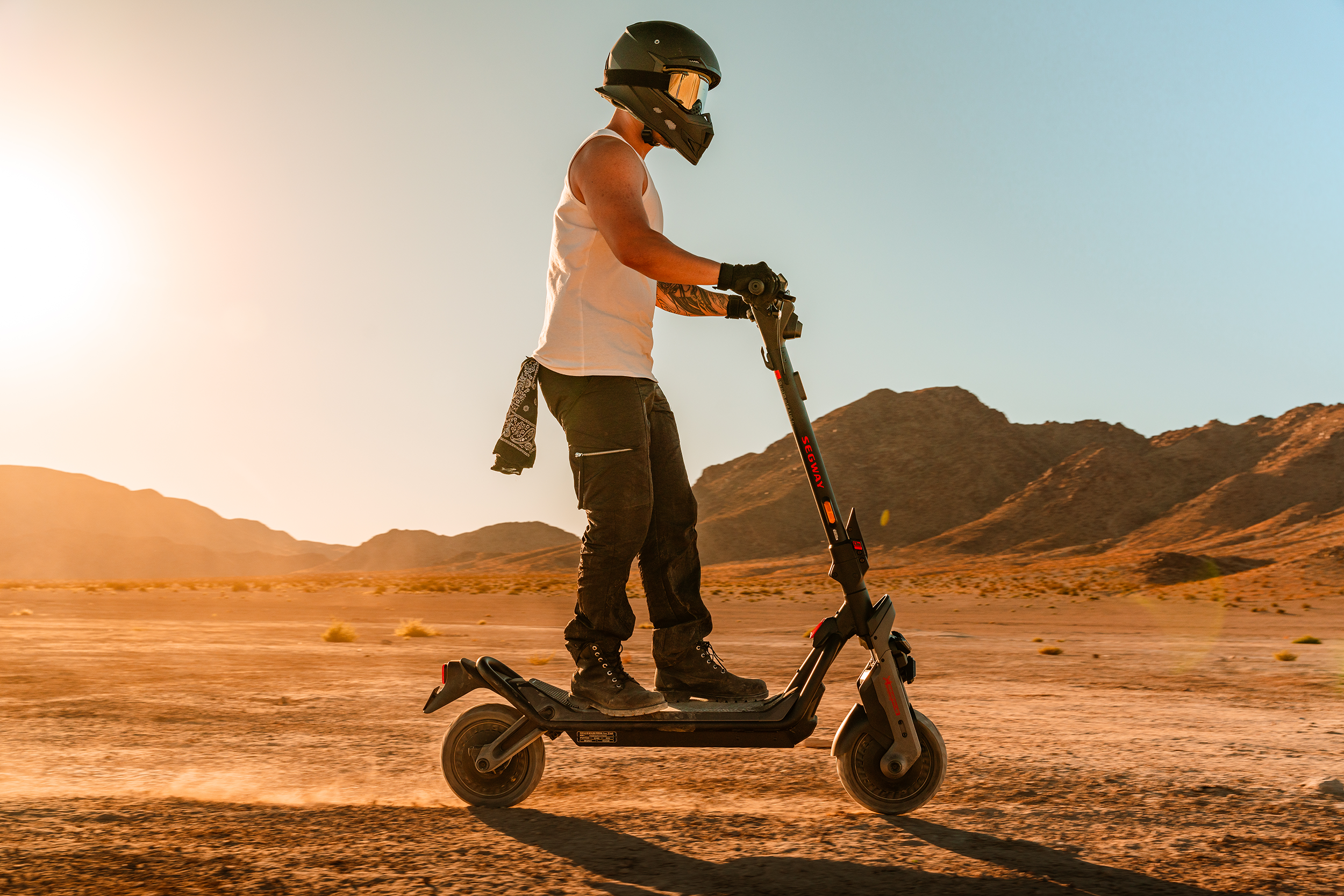 Egy sisakos személy elektromos robogóval közlekedik a sivatagban. A háttérben hegyek, tiszta égbolt látható.
