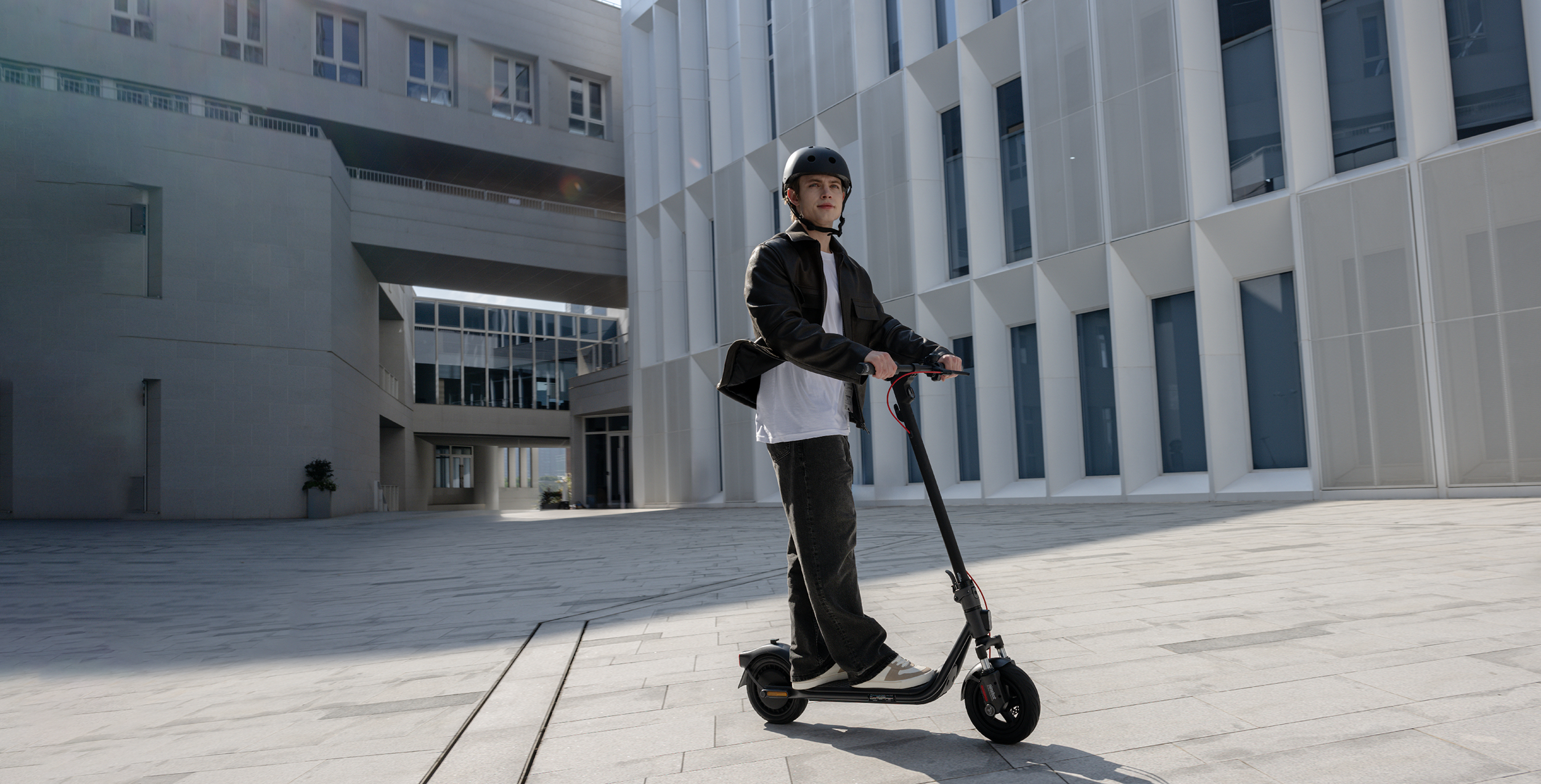 Egy személy elektromos robogón sisakot, kabátot és farmert visel. Az épületek a háttérben vannak.