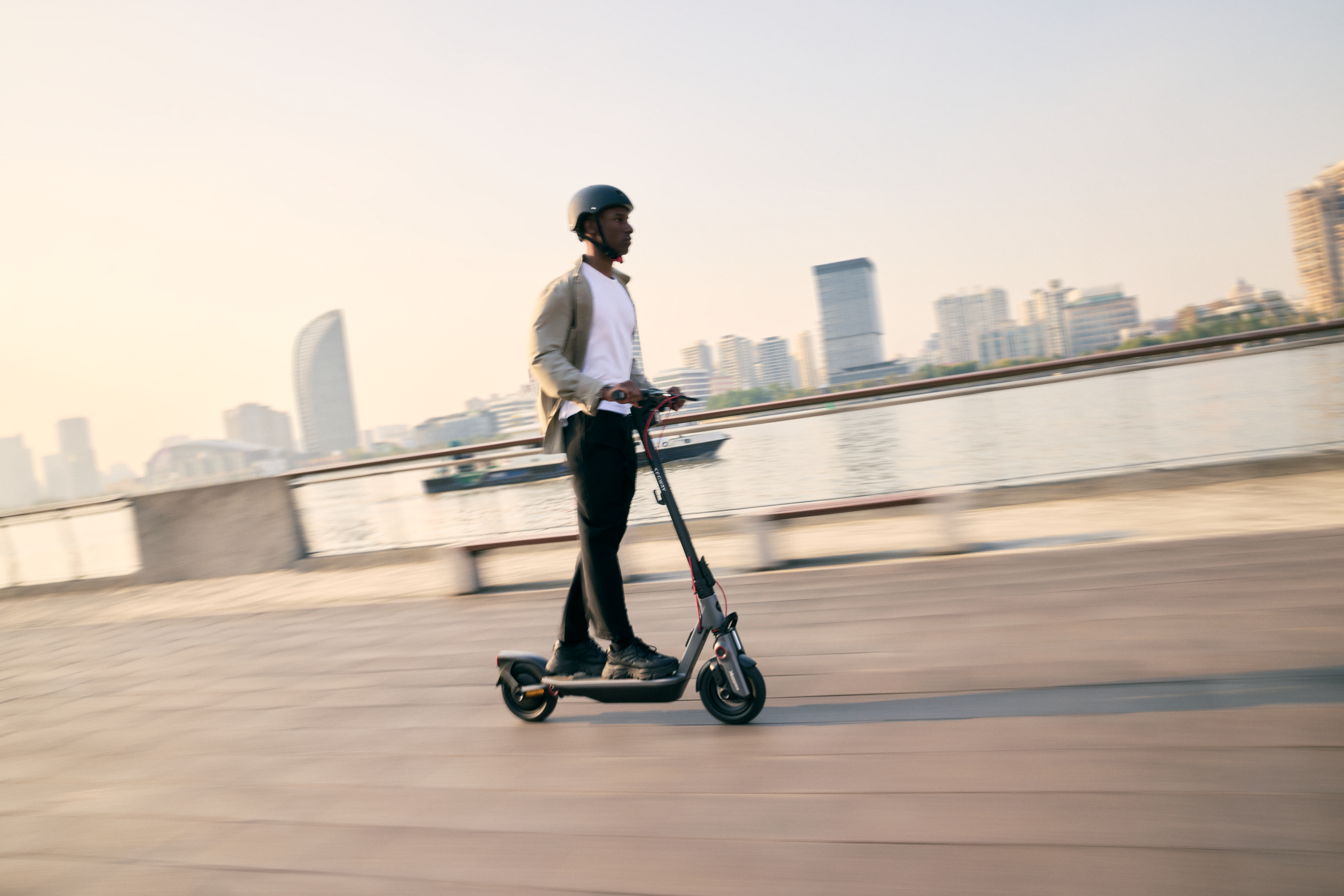 Egy férfi elektromos robogón halad egy fapályán. Sisakot és dzsekit visel. Háttérben épületek és víz láthatók.