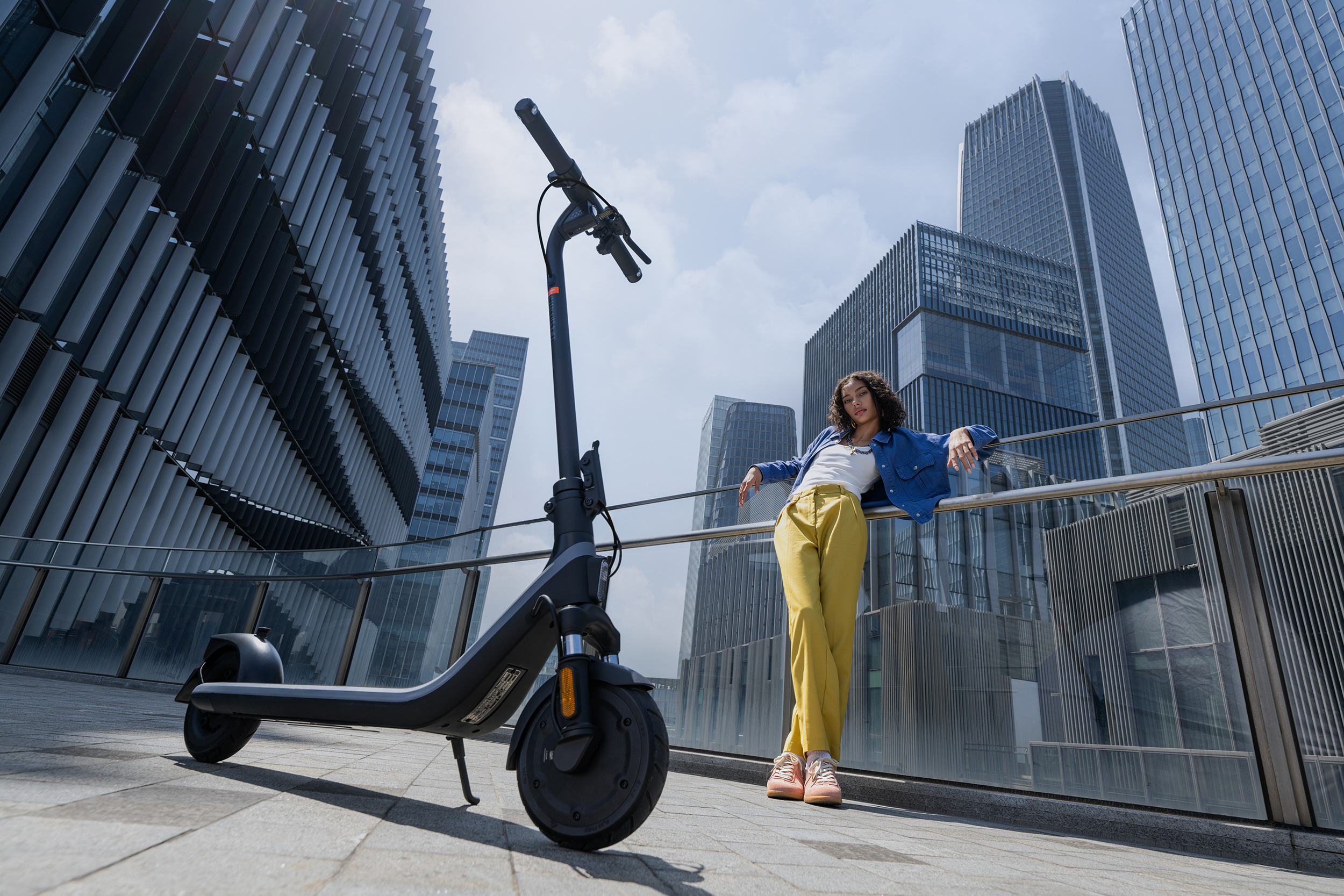 Egy nő egy elektromos roller mellett egy korlátnak támaszkodik. Felhőkarcolók a háttérben.