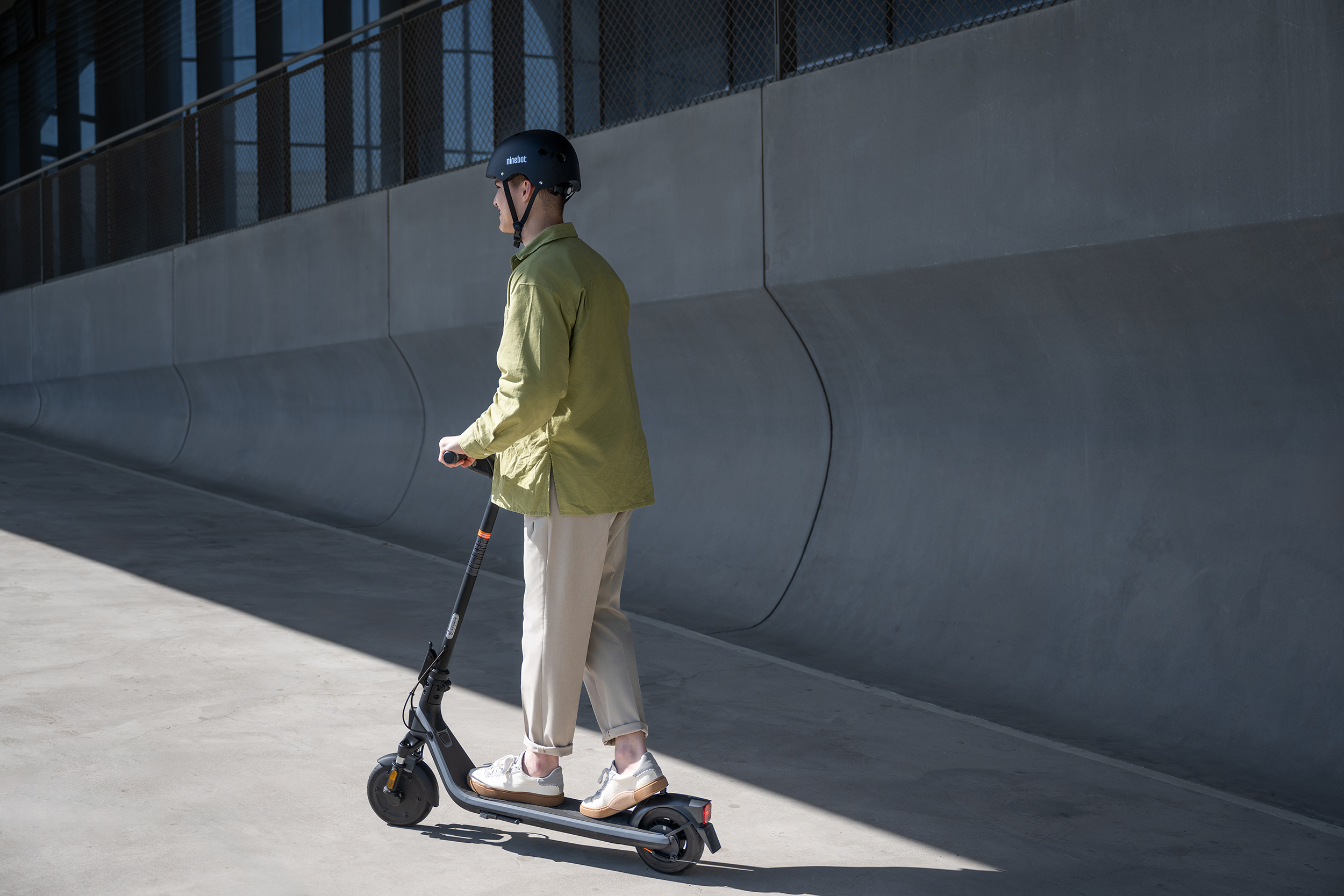 Zöld dzsekis személy elektromos robogóval közlekedik betonúton. Szürke fal és kerítés a háttérben, napos idő.