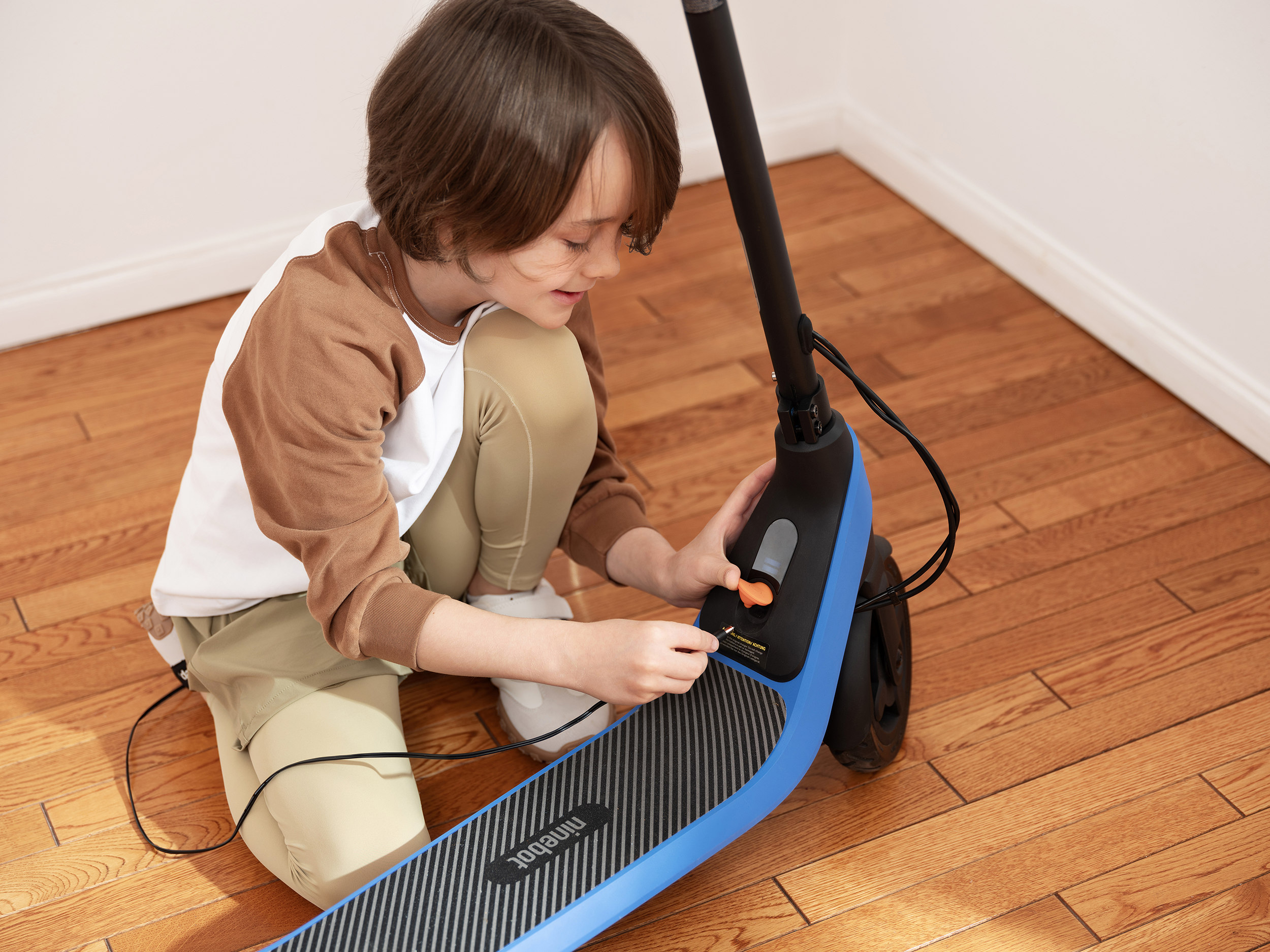 Egy gyerek térdel a fa padlón, és egy elektromos robogót állít be. A robogó kék és fekete.