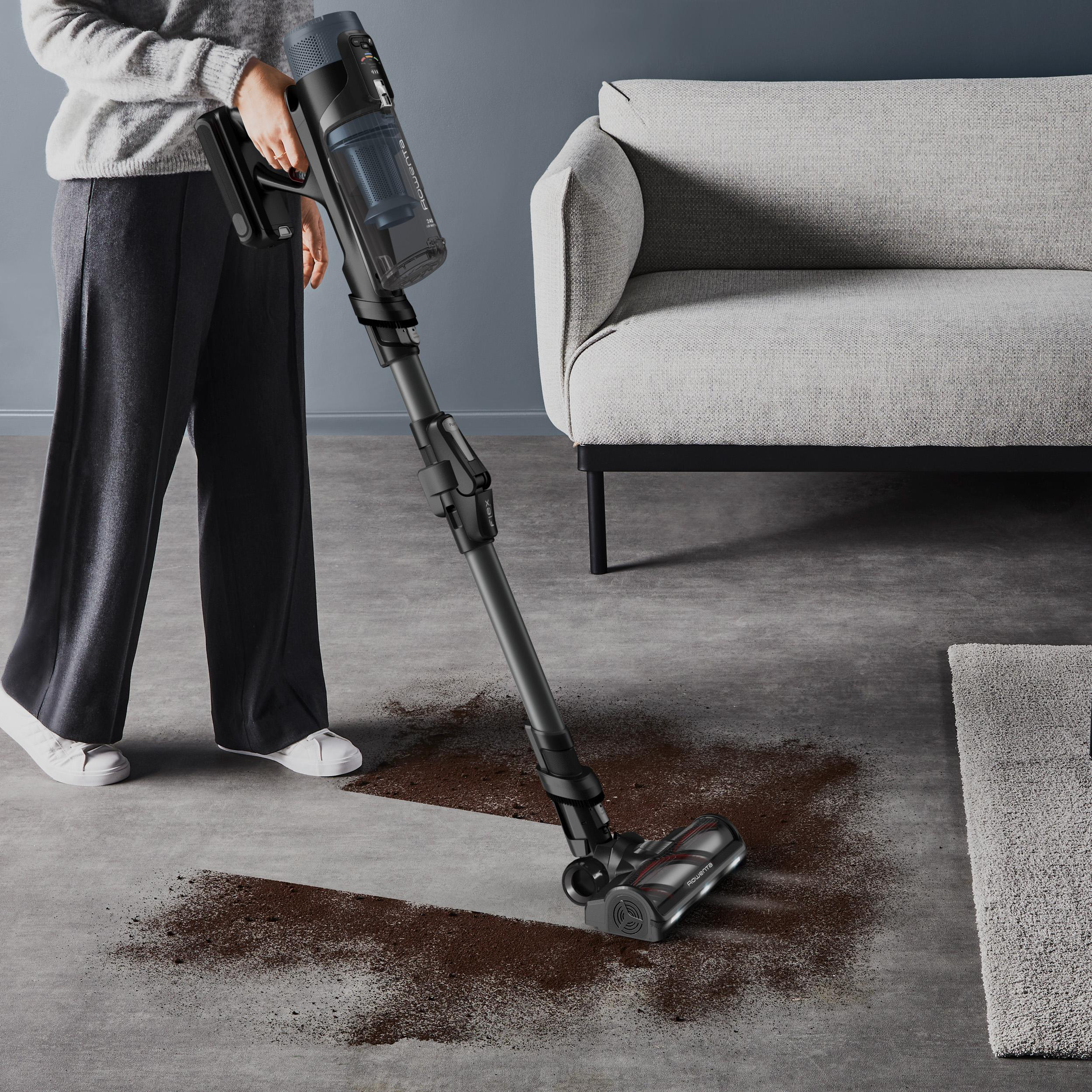Eine Person saugt Schmutz auf einem grauen Boden. Im Hintergrund ist ein graues Sofa. Der Staubsauger ist schwarz.