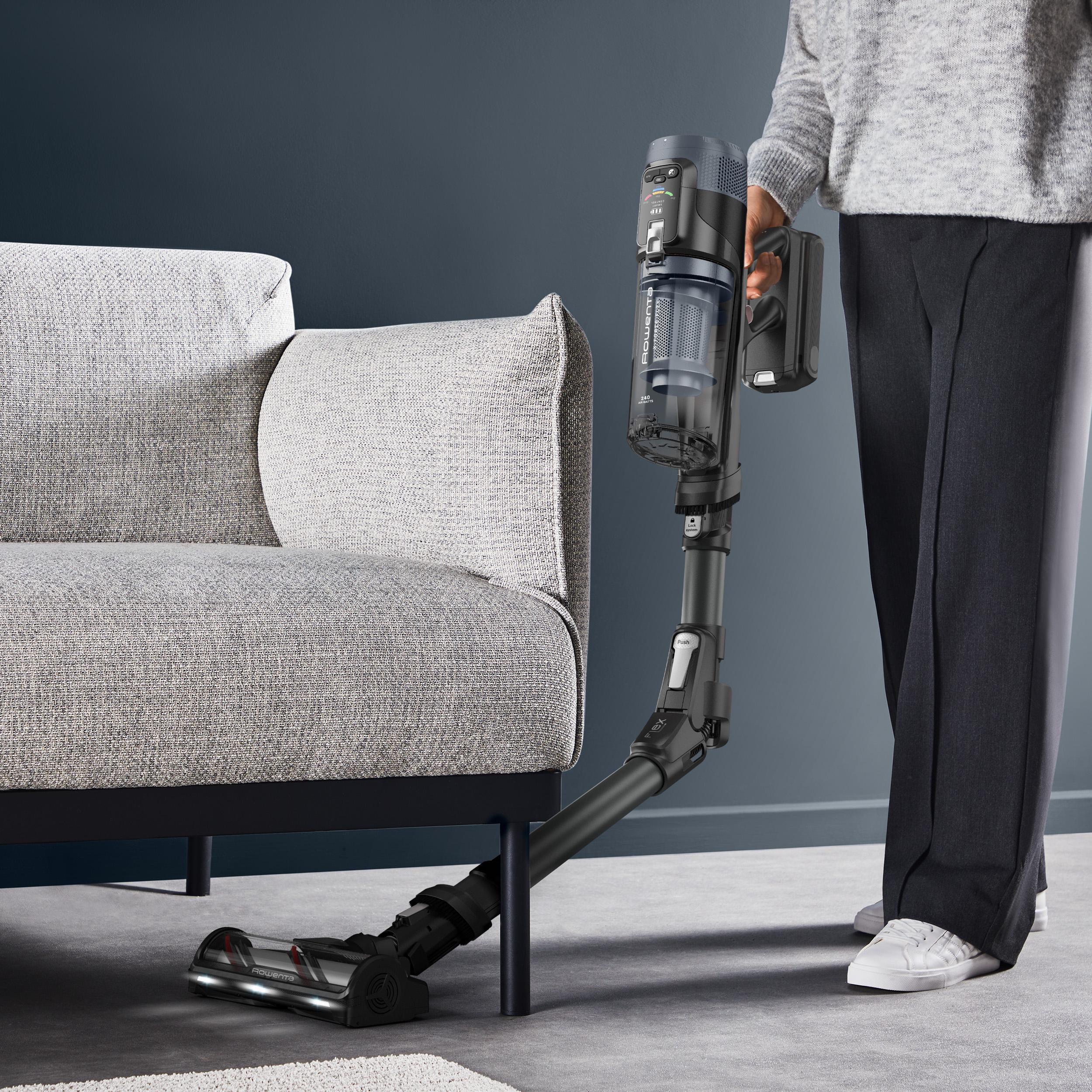 Eine Person hält einen Staubsauger in der Nähe eines Sofas. Der Staubsauger ist schwarz und grau. Die Wand ist dunkelblau.