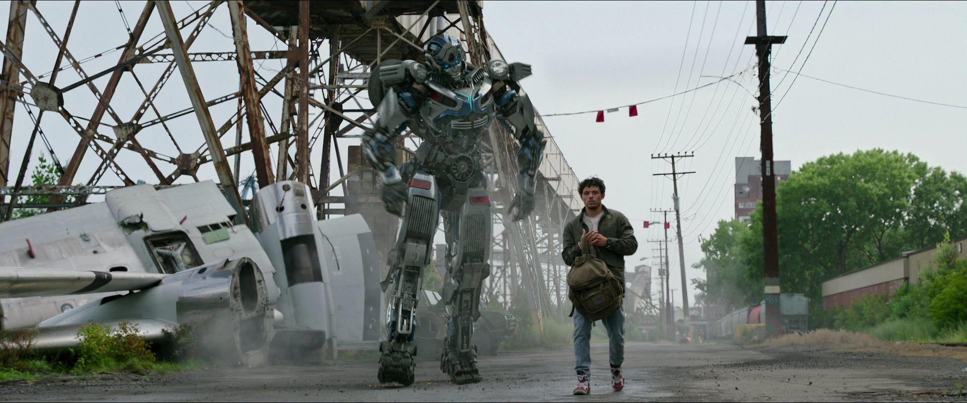 Ein Mann geht auf die Kamera zu. Ein großer Roboter steht hinter ihm. Ein beschädigtes Flugzeug ist links. Die Szene ist im Freien.