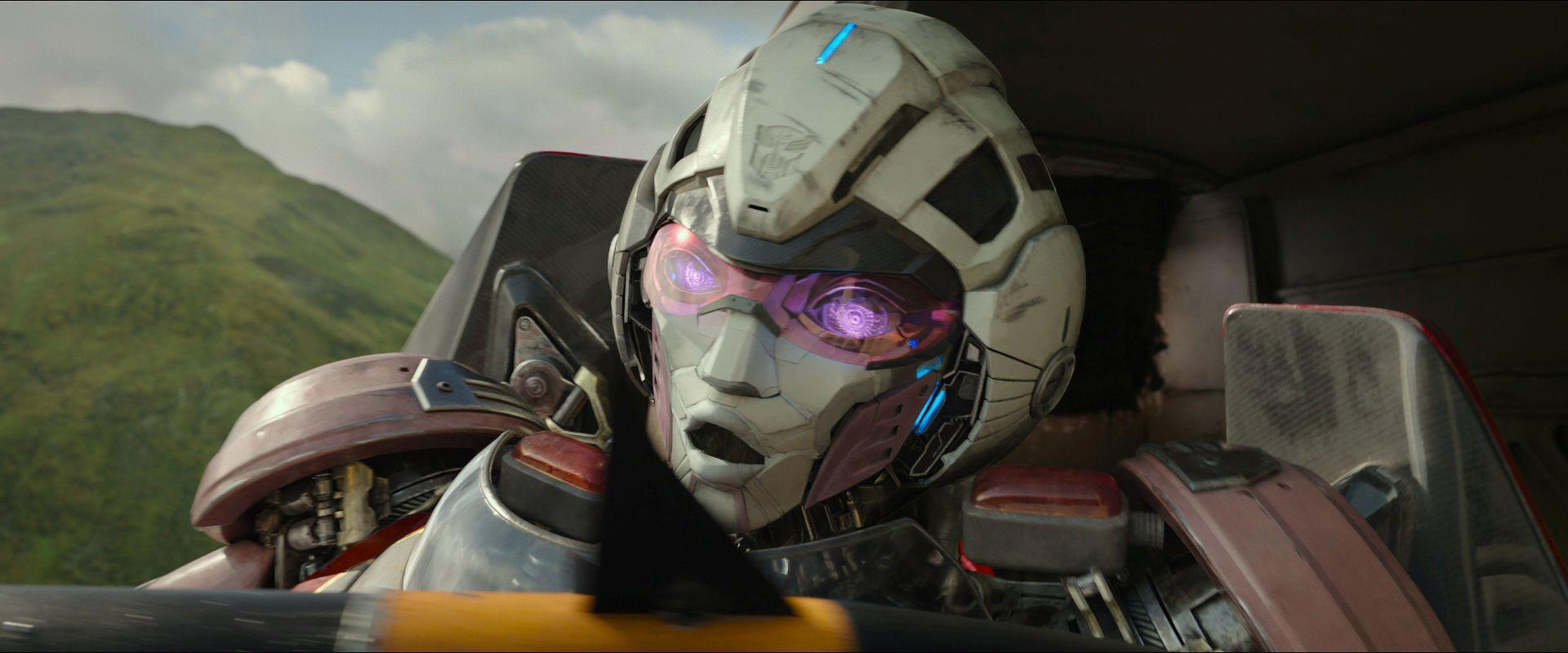 Ein Robotergesicht mit rosa Augen. Es ist hell und hat dunkle Details. Es befindet sich in einem Fahrzeug vor Bergkulisse.