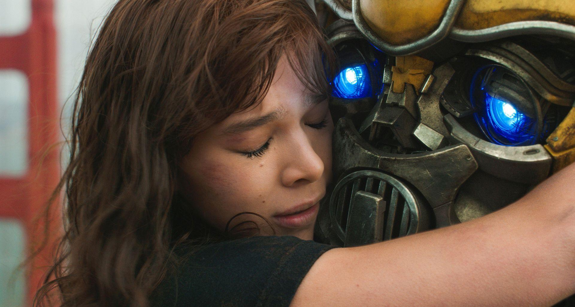 Eine junge Frau umarmt einen Transformer. Ihre Augen sind geschlossen. Der Roboter hat gelbe und graue Metallteile mit blau leuchtenden Augen.