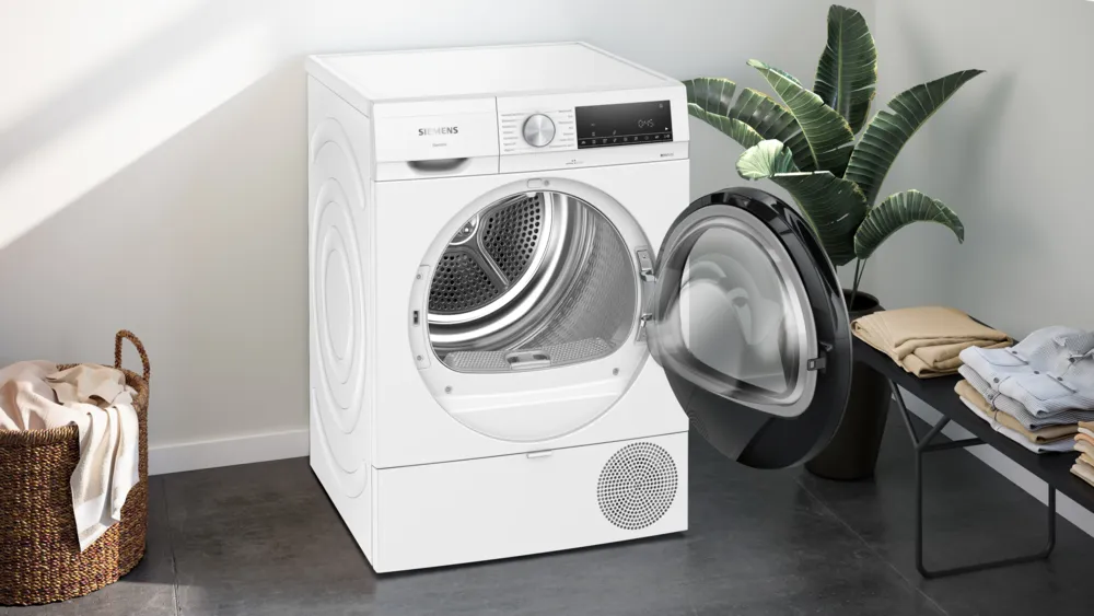 Un sèche-linge Siemens blanc avec une porte ouverte. Un panier de vêtements à gauche. Un banc avec des vêtements pliés à droite.