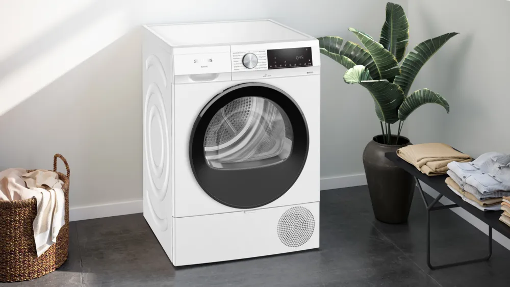 Un sèche-linge Siemens blanc se trouve près d'une plante en pot, des vêtements sont empilés sur une table noire et un panier en osier à gauche.
