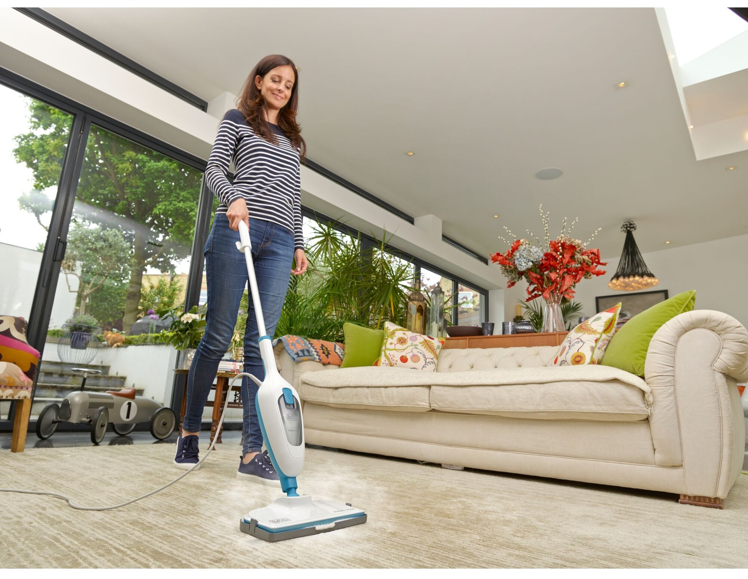 Une femme nettoie un tapis avec un nettoyeur vapeur. Elle porte une chemise rayée et un jean. Un canapé et une fenêtre sont derrière elle.