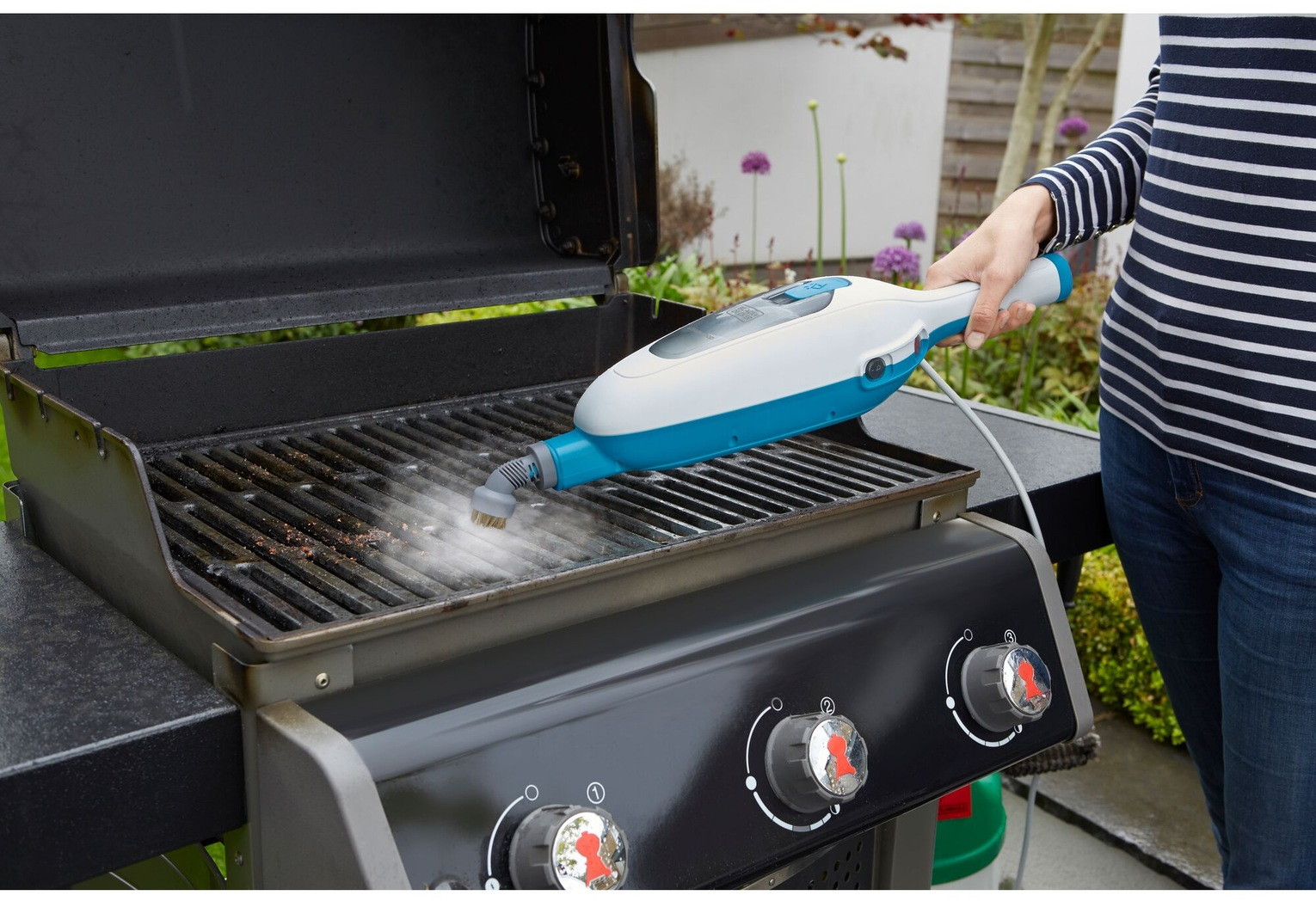 Une personne nettoie un barbecue avec un nettoyeur vapeur. Le barbecue est noir, et le nettoyeur blanc et bleu. De la vapeur monte.