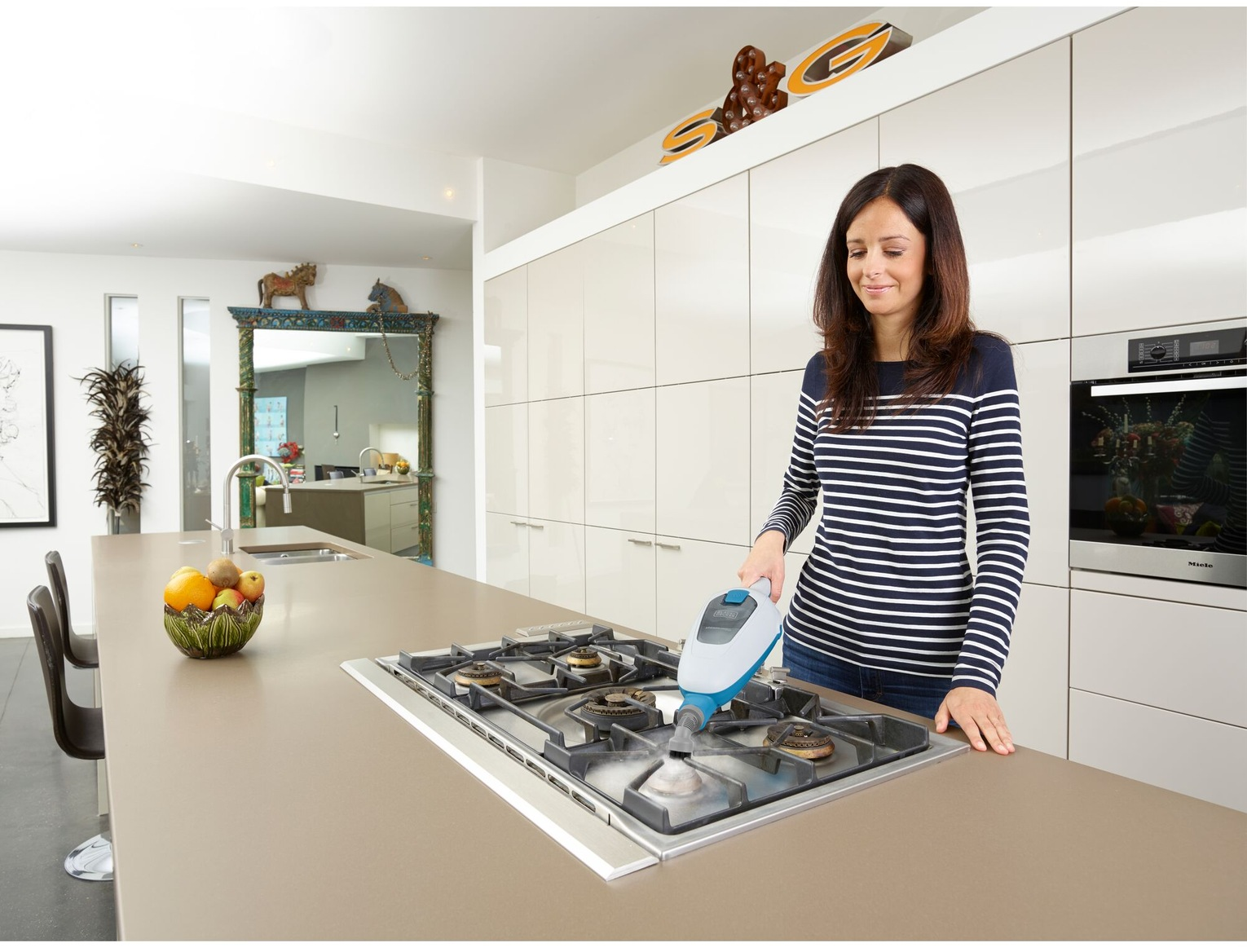 Femme nettoyant une cuisinière à gaz avec un nettoyeur vapeur. Cuisine avec meubles blancs, coupe de fruits et miroir en arrière-plan.