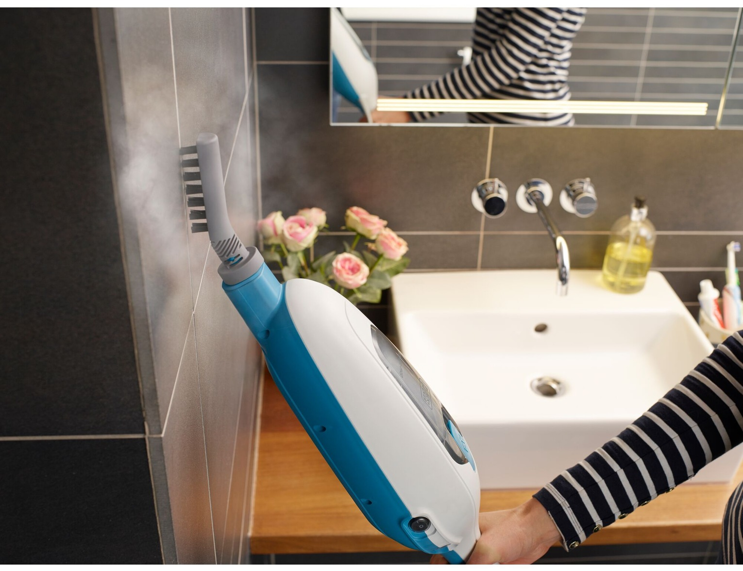 Une personne utilise un nettoyeur vapeur bleu et blanc sur un mur de salle de bain carrelé. Un miroir et un lavabo sont en arrière-plan.