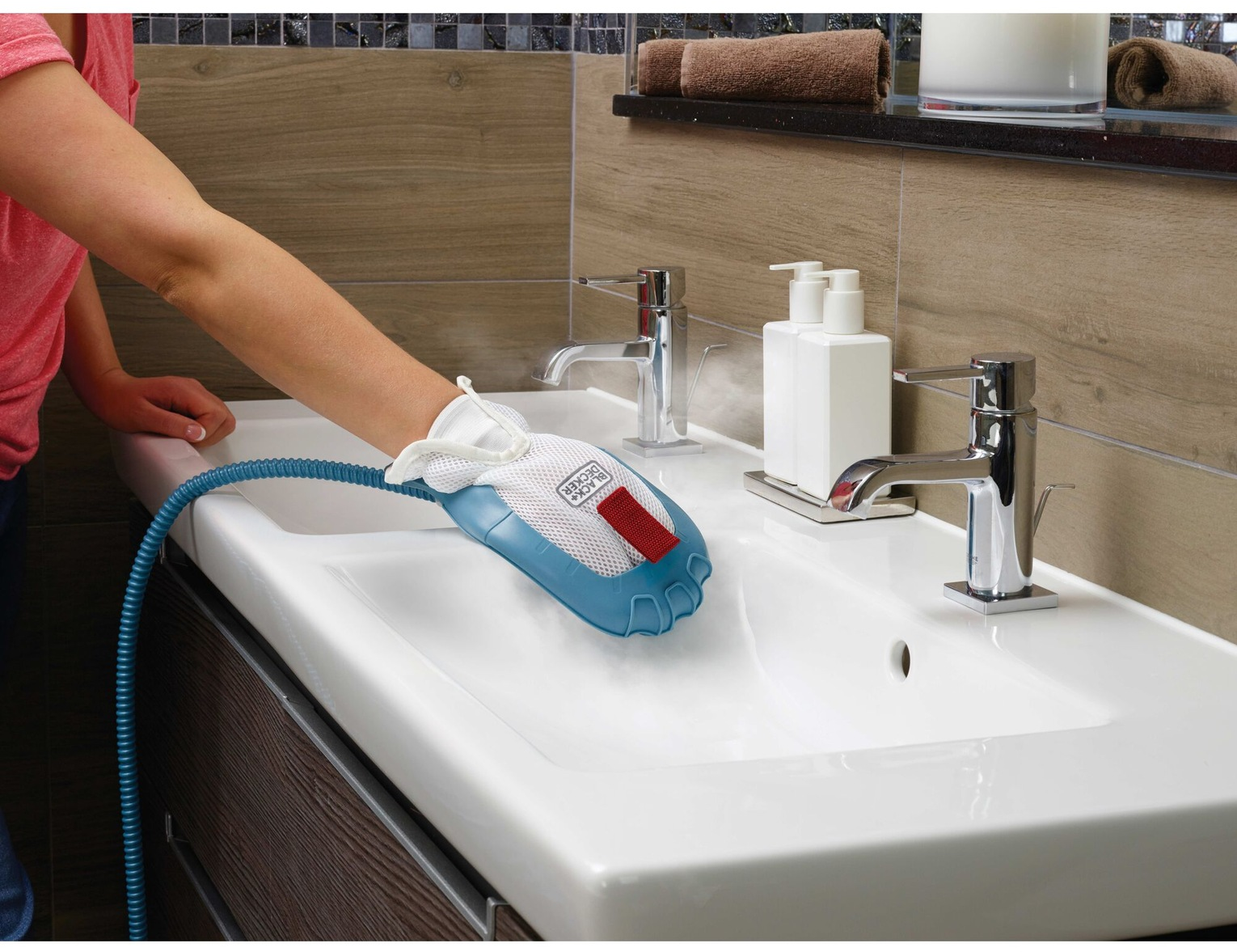 Una persona usa un vaporizzatore su un lavandino bianco. Viene utilizzato uno strumento di pulizia blu e bianco. Sono visibili anche rubinetti e dispenser di sapone.