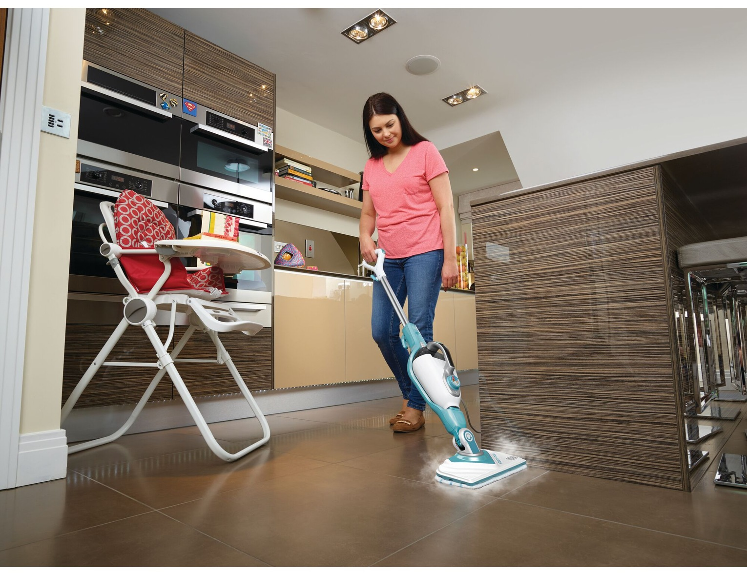 Una donna pulisce il pavimento della cucina con un pulitore a vapore. Un seggiolone è vicino. Nel retro ci sono armadi e elettrodomestici.