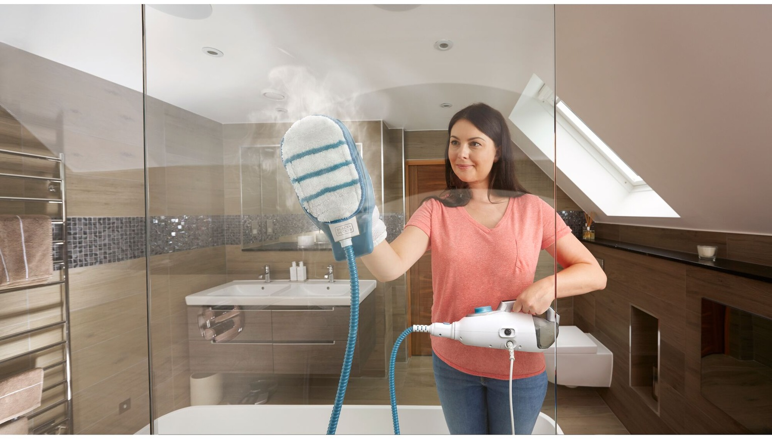 Donna che pulisce una porta della doccia in vetro con un pulitore a vapore. Il bagno ha un lavandino, un WC e un portasciugamani.