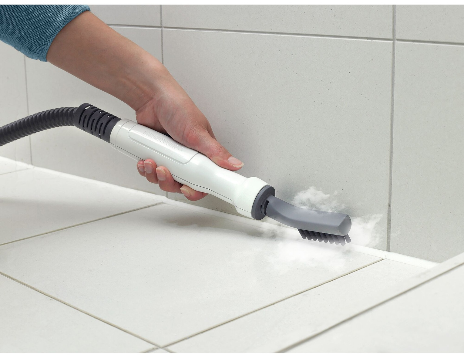 Una mano tiene un pulitore a vapore bianco e grigio, pulendo piastrelle bianche. Il vapore esce dalla testina.