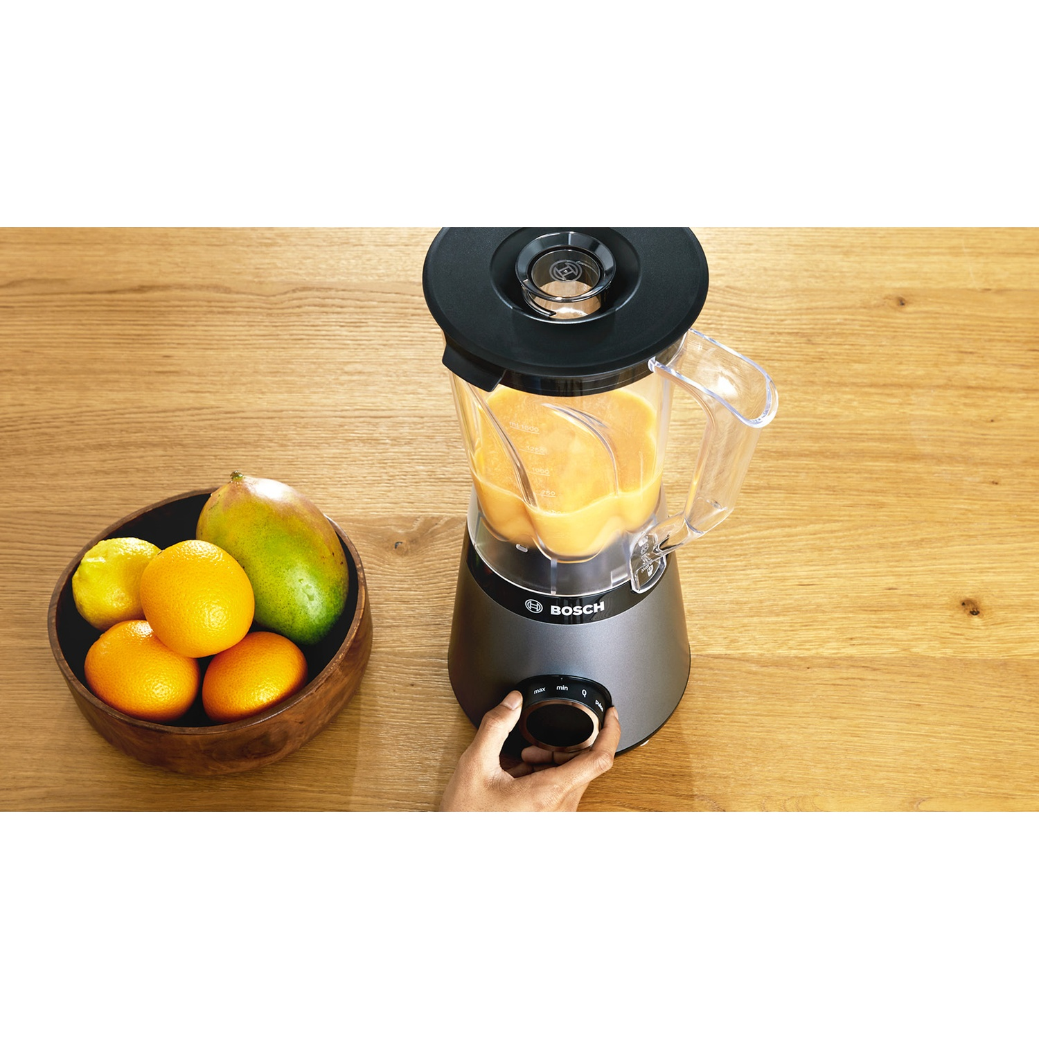 Un frullatore con liquido arancione è su un tavolo di legno. Una ciotola di frutta, tra cui arance e mango, è a sinistra.