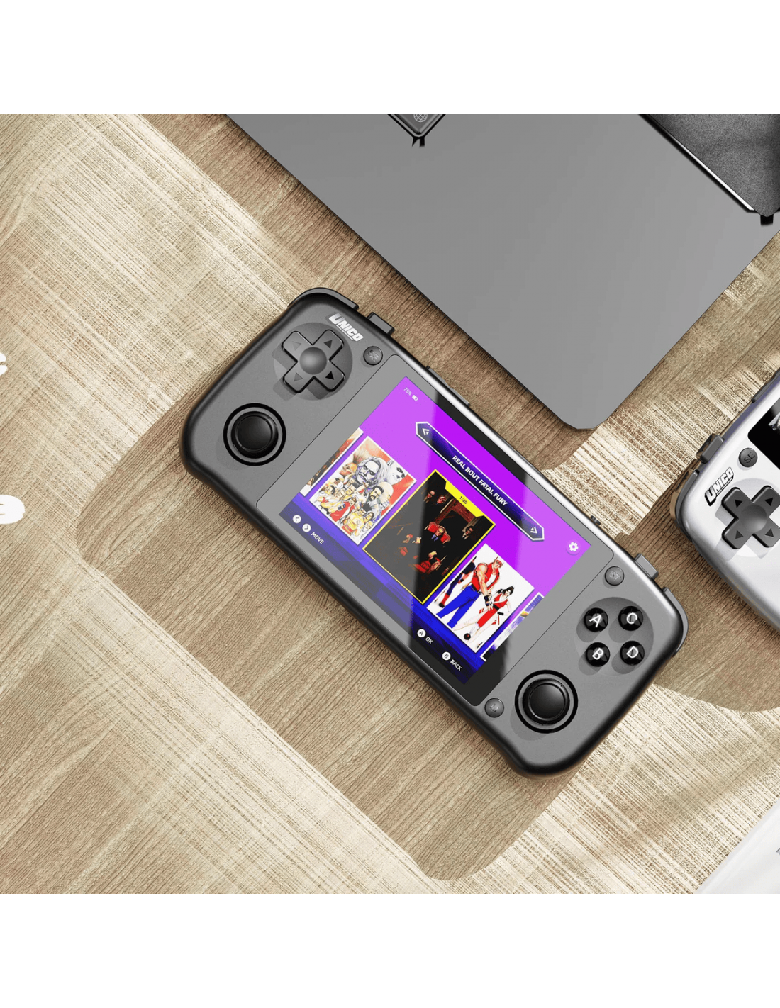Consola de juegos portátil negra con pantalla mostrando opciones de juego, sobre una mesa de madera. Un portátil es parcialmente visible.