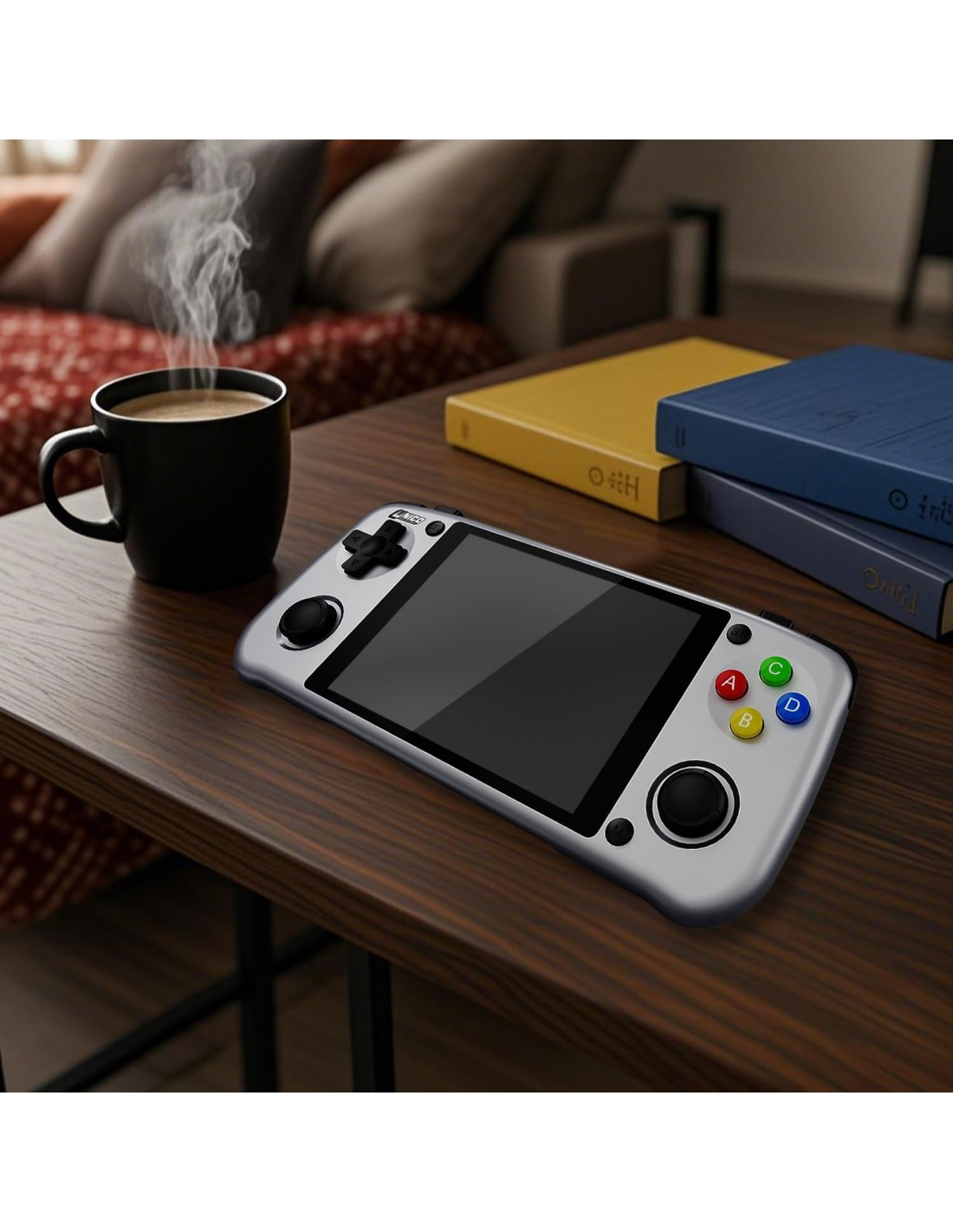 Una consola de juegos portátil plateada sobre una mesa de madera. Una taza de café humeante y libros en el fondo. Sofá visible.