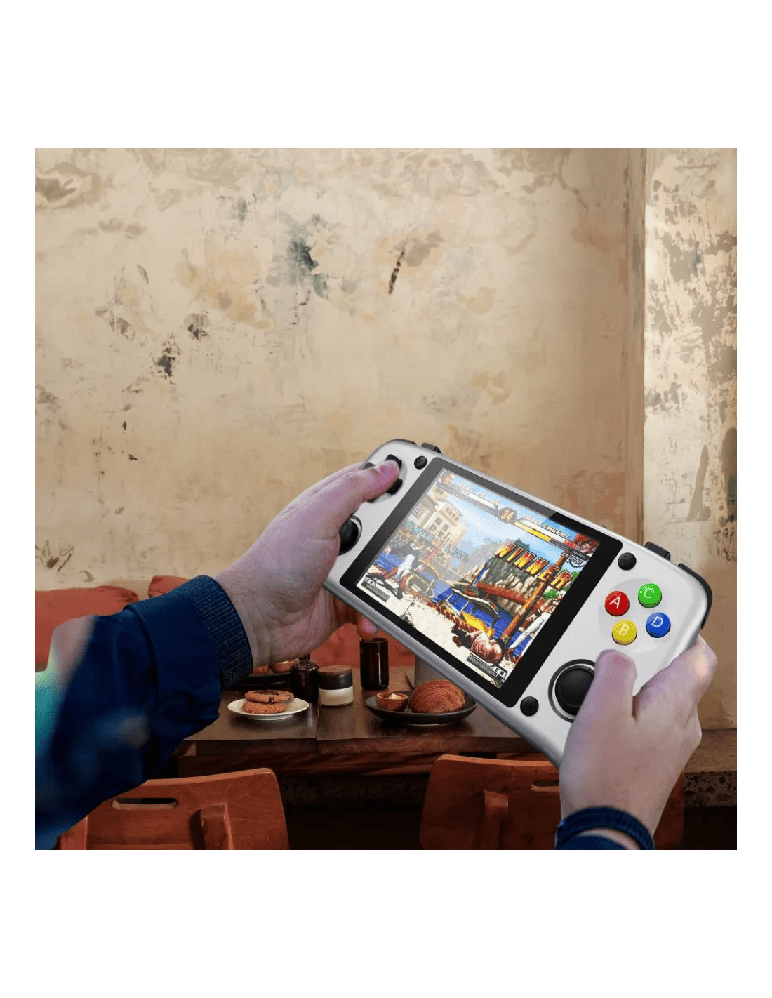 Manos sosteniendo una consola de juegos portátil blanca. Se muestra un juego. Mesa con comida en primer plano, una pared en el fondo.